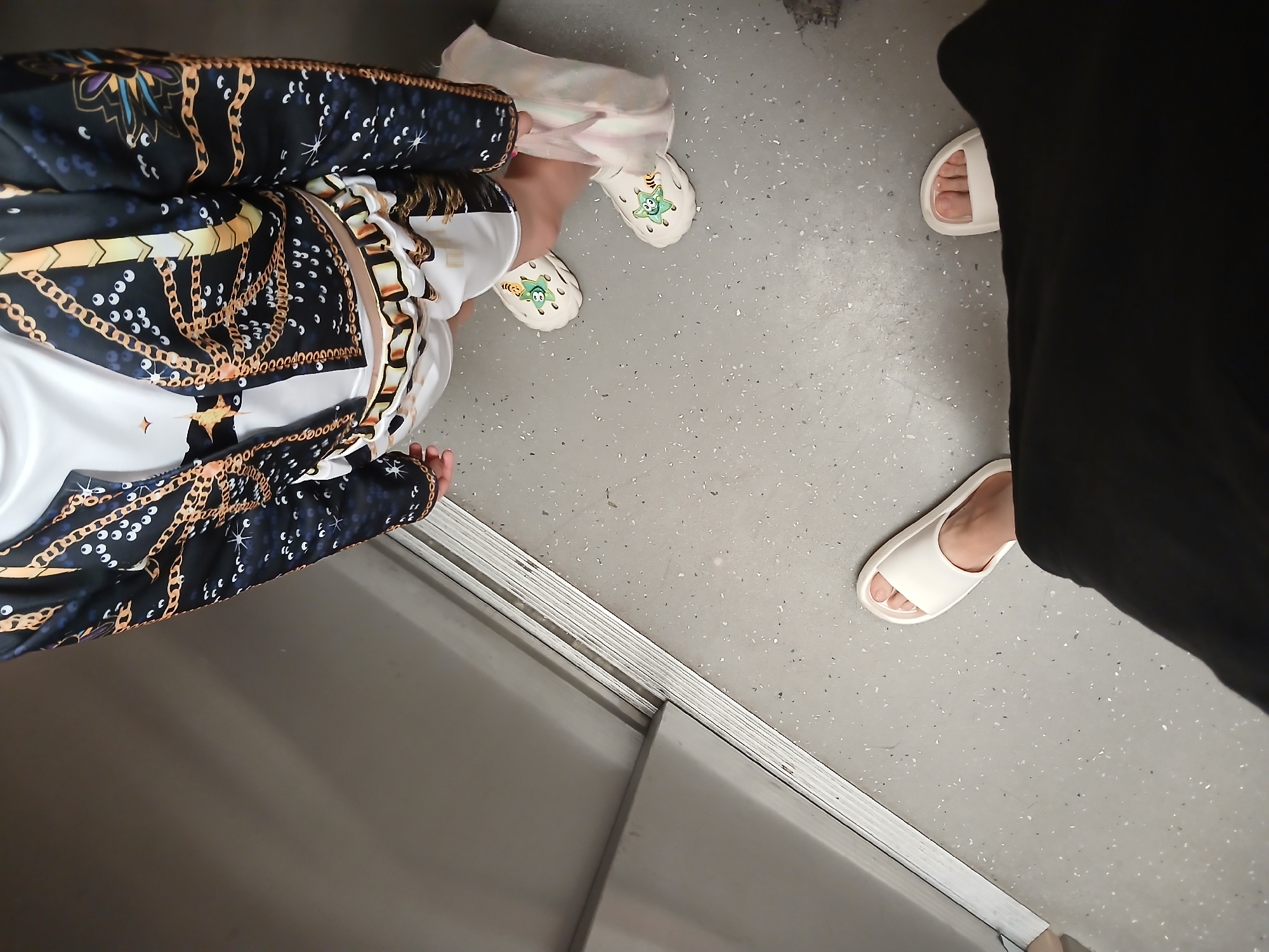 The image shows a close-up view of a person's legs and feet inside what appears to be an elevator. The person is wearing a black jacket with a gold chain pattern, white shorts with a similar pattern, and white slides. One foot is partially covered by a light-colored fabric, possibly a towel or garment. Another person's legs and feet, wearing white slides and black clothing, are visible in the foreground. The floor is a speckled gray surface, and a metallic sliding door is visible on the left. The image provides no information or context to suggest a location in Yekaterinburg, Russia.