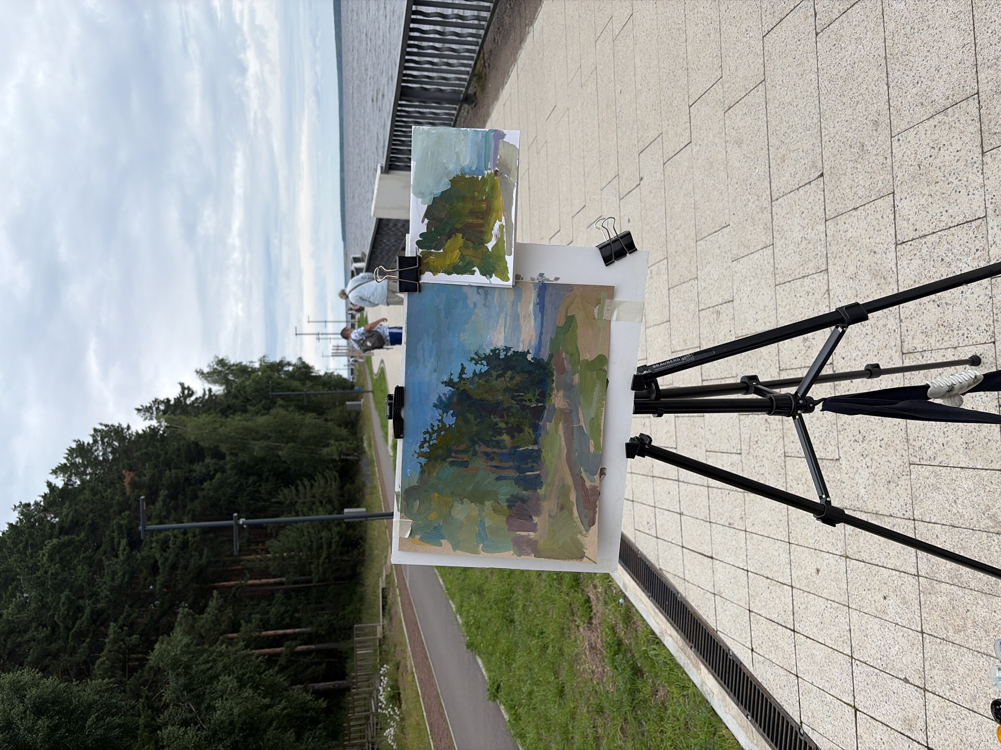 A plein air painting setup is displayed on a tiled promenade next to a body of water. The foreground is dominated by a black tripod supporting two paintings. The larger painting, on the left, depicts a forest scene with a blue sky and water. To its right, a smaller study shows trees and a cloudy sky. Two people are walking away from the viewer along the promenade, towards the right of the frame. Beyond them, a black metal railing runs along the edge of the water, which appears to be a river or lake. On the left side of the promenade, a paved path is visible, bordered by a grassy area and a dense forest. Streetlights are positioned along the promenade. The sky is overcast with clouds. The location is the embankment in the city of Chaikovsky, Russia, a district situated along the Kama River.