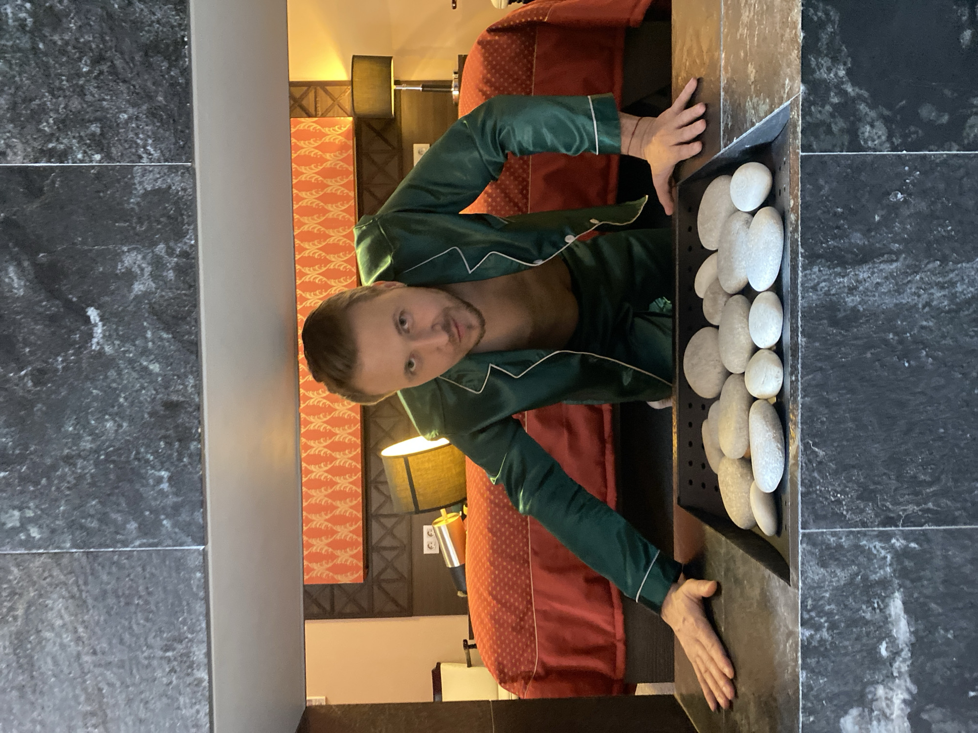 A fair-skinned man in dark green pajamas with white trim is looking at the camera. He is behind a fireplace in a hotel room, possibly in Yekaterinburg, Russia, given the Russian-language text in the image description. The fireplace is a dark, vertical rectangle with a black grate holding a collection of smooth, white stones. The man’s hands rest on the fireplace mantel.
Behind the man and fireplace is a hotel room. The bed has a red comforter. On the wall behind the bed is an orange patterned wallpaper. There is a lamp on the nightstand. The room has dark, wooden trim. To the left of the fireplace is a wall tiled with dark gray stone.