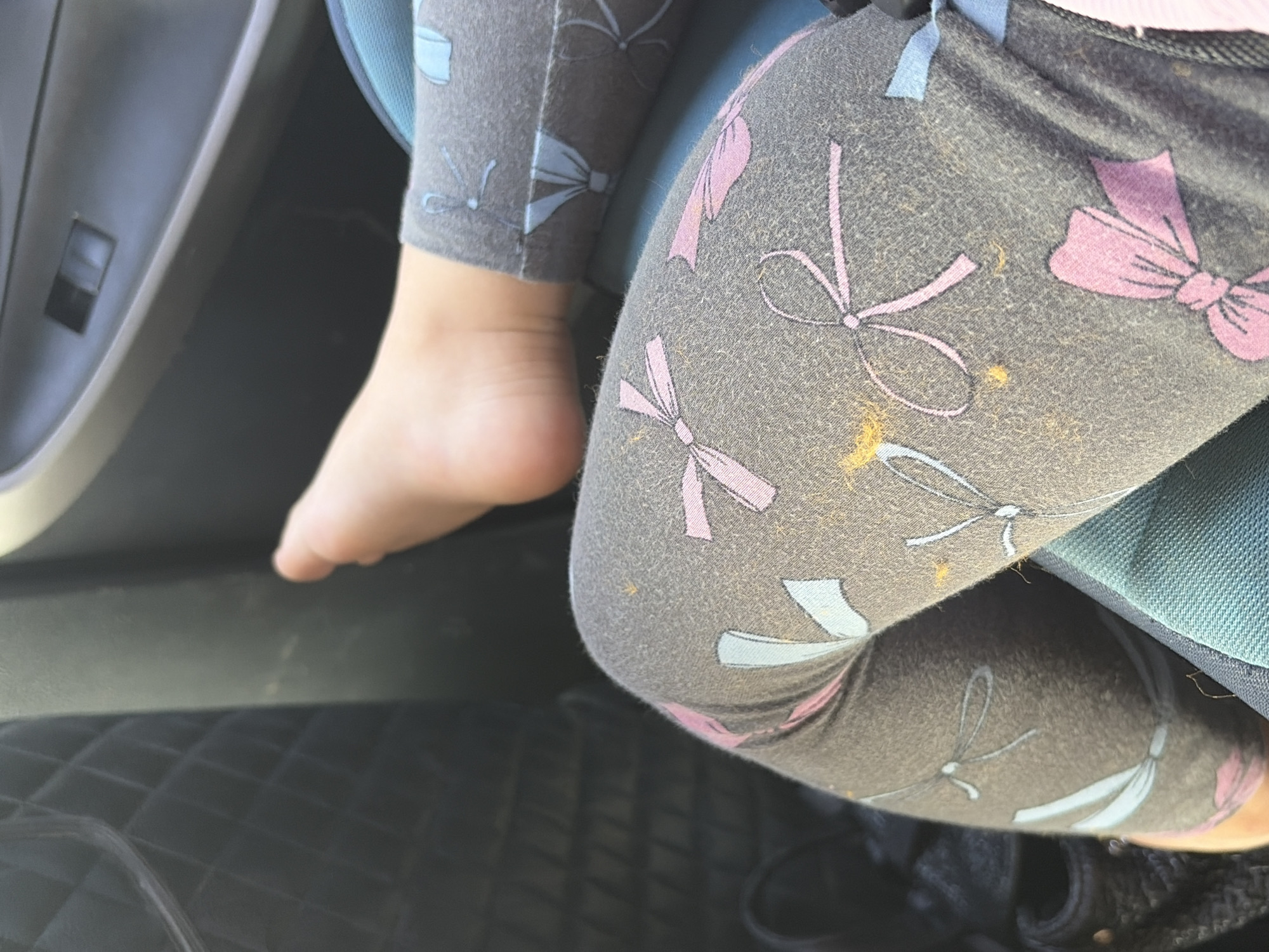 The image shows a close-up view of a child's bare foot and legs, wearing grey leggings with a pattern of pink and light blue bows. The child is seated in a car, as indicated by the car door panel visible in the upper right corner, and the quilted texture of a car seat on the left. The background is out of focus, but suggests an indoor or vehicular environment. There are no discernible landmarks or contextual clues to indicate the specific location within Rostov-on-Don, Russia.