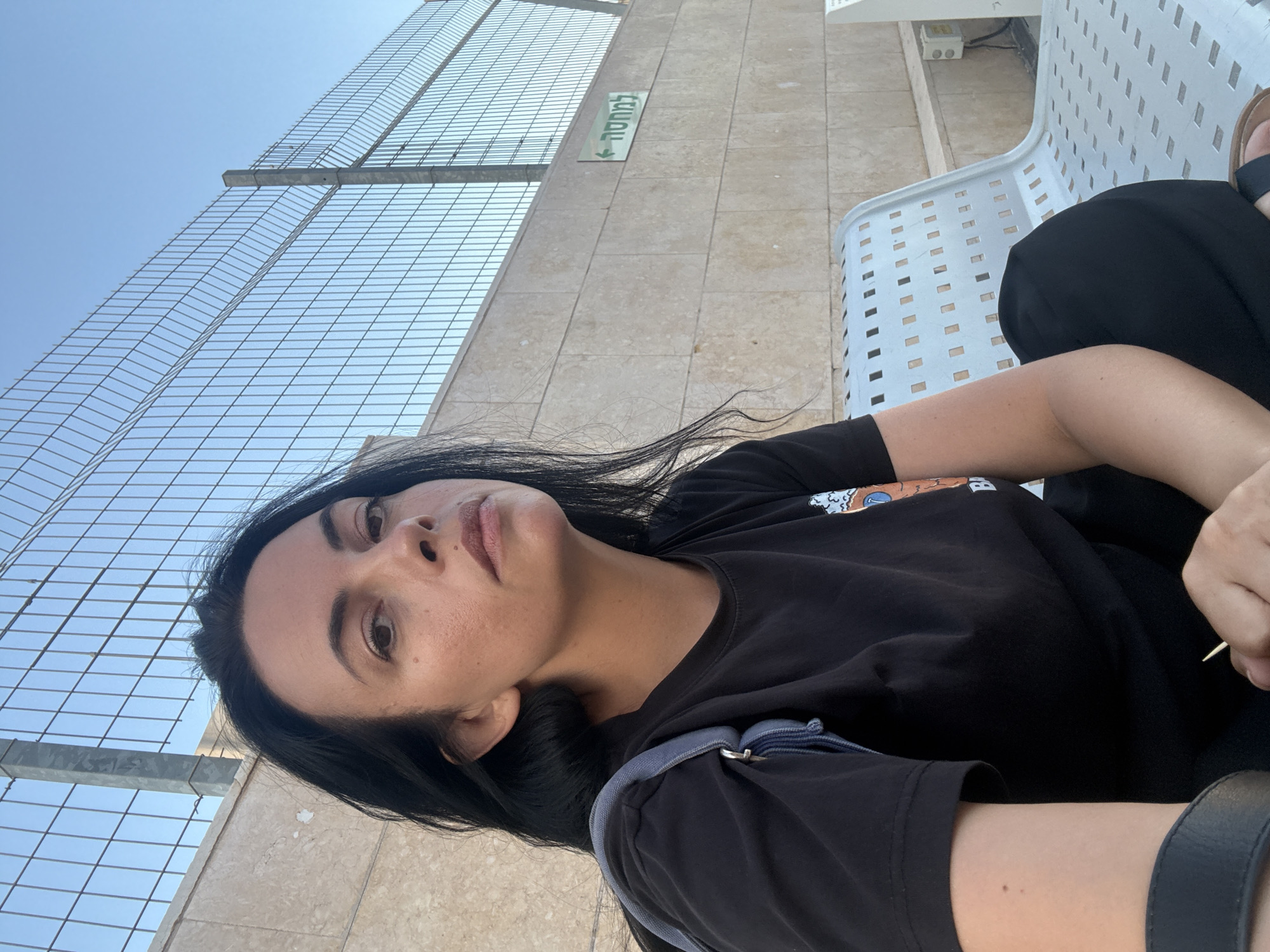 The image shows a woman sitting on a metal bench in Hod HaSharon, Israel. She is wearing a black t-shirt and black pants. A light blue bag is slung over her shoulder. The background consists of a stone wall with a green sign pointing to the left, and a wire fence with a clear sky beyond. The bench has a perforated design.