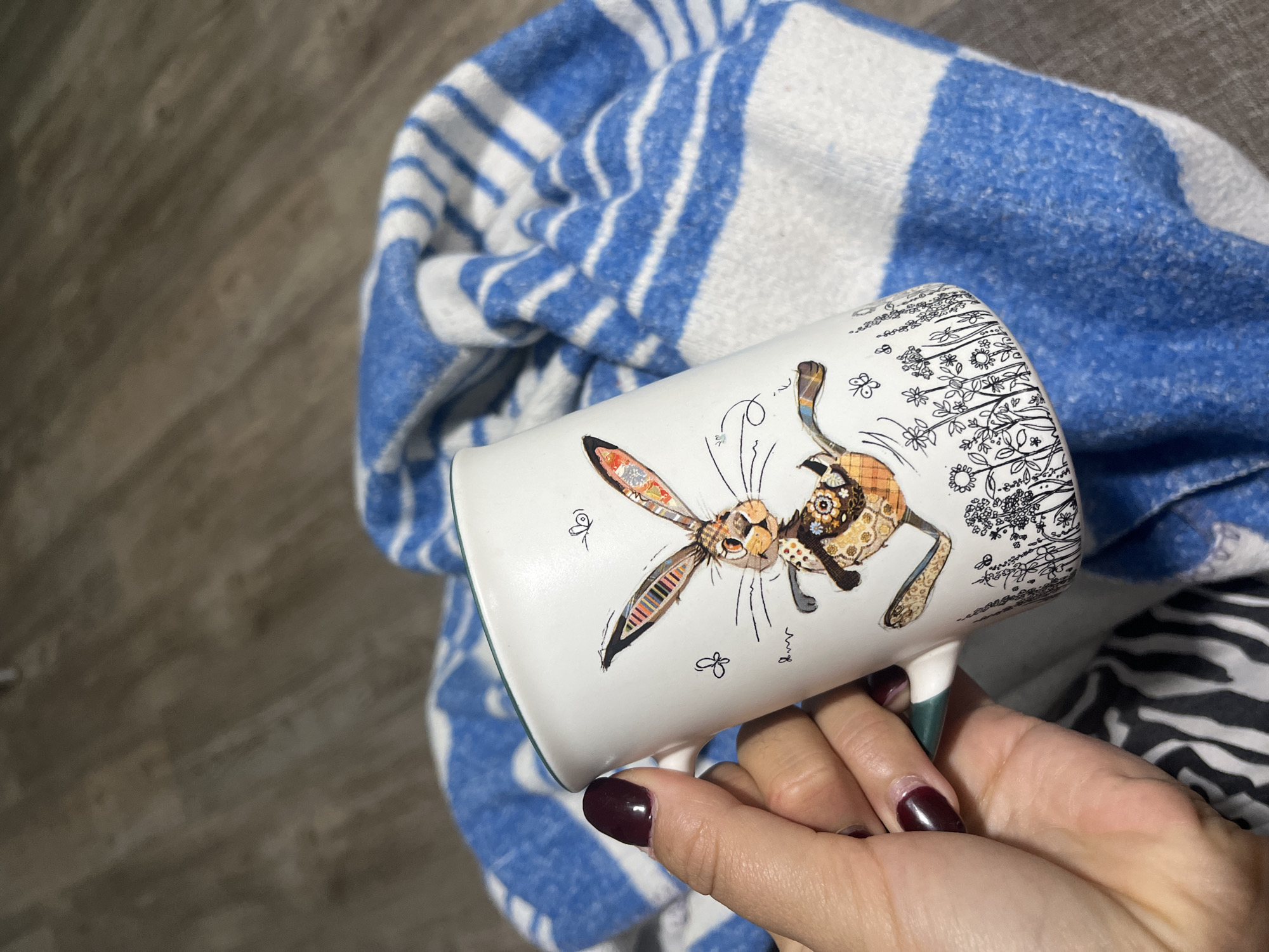 A hand with dark red nail polish holds a white mug decorated with a whimsical rabbit and small butterflies, set against a blue and white striped blanket. The mug has a green rim. The background appears to be a wooden floor. The scene is indoors. The mug and blanket are the primary subjects. There are no people visible in the image, only a hand holding the mug.
