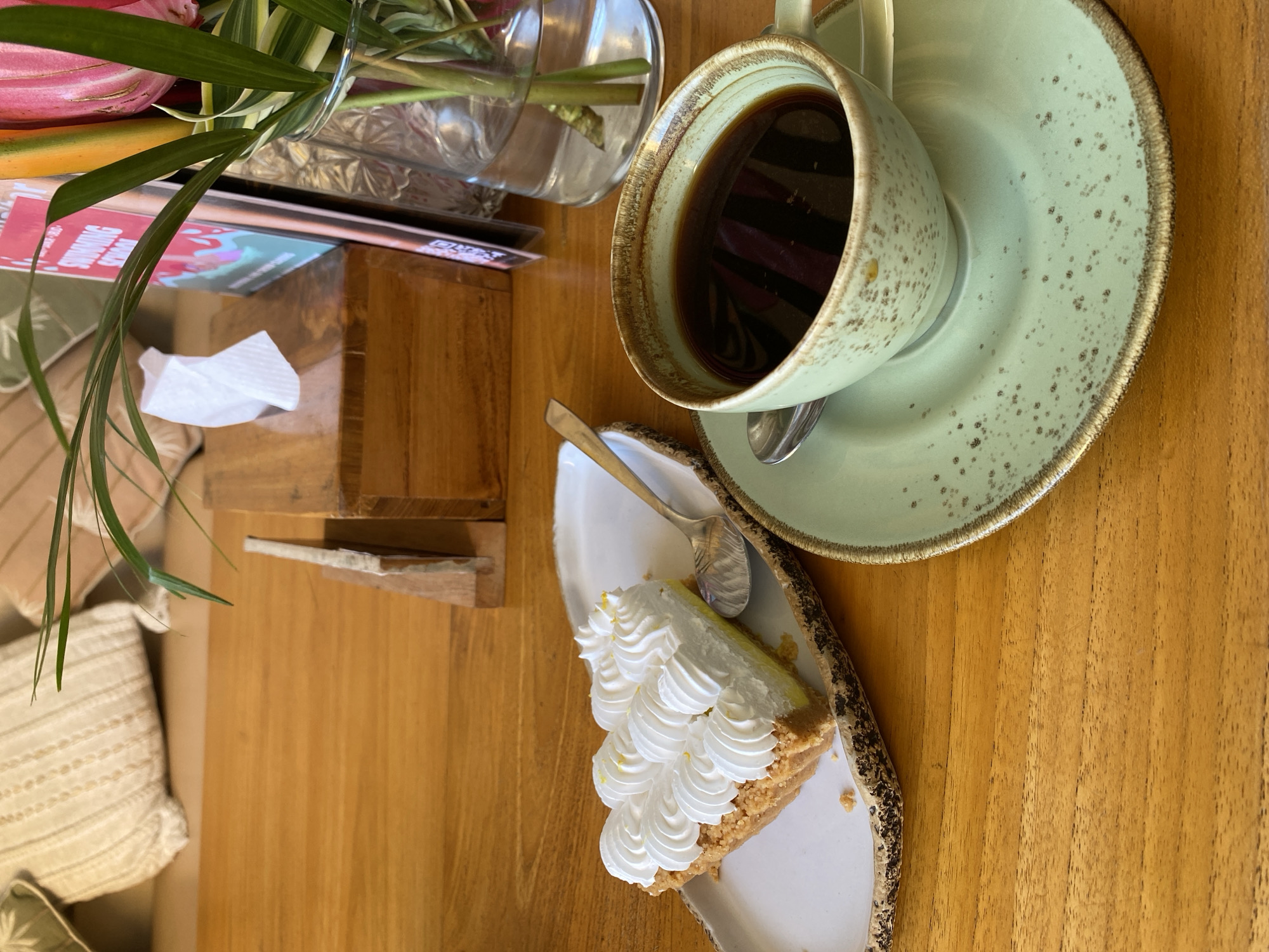 A high-angle, indoor shot reveals a light wooden table arranged with various items. A green cup of black coffee on a matching saucer is positioned on the upper right of the frame. Next to it, a slice of pie topped with decorative white meringue sits on an irregularly shaped plate. A small spoon rests on the plate near the pie. Adjacent to the pie, there is a small wooden structure with a small white napkin. A vase holding flowers is placed in the upper left corner of the scene. A menu card, partially visible behind the vase, adds to the composition. The shot was taken in Kuta Selatan, Indonesia.