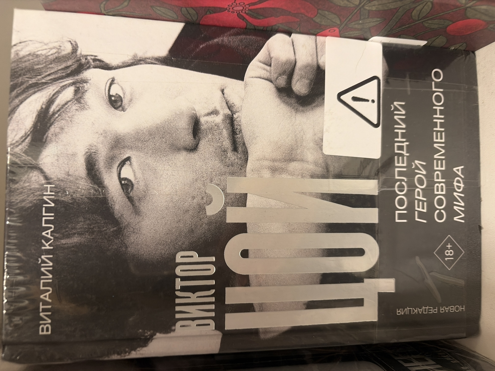The media file shows a book cover, likely a biography, oriented vertically and rotated ninety degrees clockwise. The cover features a high-contrast, black and white portrait of a man, possibly Viktor Tsoy, looking upwards with a thoughtful expression. Part of a clenched fist is visible on the right side of the man's face.
Prominently displayed in large, silver, block letters across the lower right portion of the cover is the name "ЦОЙ" (Tsoy). Below this, in smaller silver text, is "ВИКТОР" (Viktor), indicating the full name Viktor Tsoy. The author's name, "ВИТАЛИЙ КАЛГИН" (Vitaliy Kalgin), is printed vertically along the left edge of the cover in white text. Along the right edge, also in white vertical text, is the subtitle: "ПОСЛЕДНИЙ ГЕРОЙ СОВРЕМЕННОГО МИФА" (The Last Hero of a Modern Myth). A small, inverted text in the bottom right corner reads "НОВАЯ РЕДАКЦИЯ" (New Edition).
A white rectangular sticker with a black triangular warning symbol (exclamation mark inside) is placed over the man's right hand and the book's title, partially obscuring the image. Below the subtitle, a black square with rounded corners contains the age restriction "18+". The book cover appears to be wrapped in plastic film, and behind it, a red and green floral pattern is partially visible, suggesting it's resting on another object or surface.