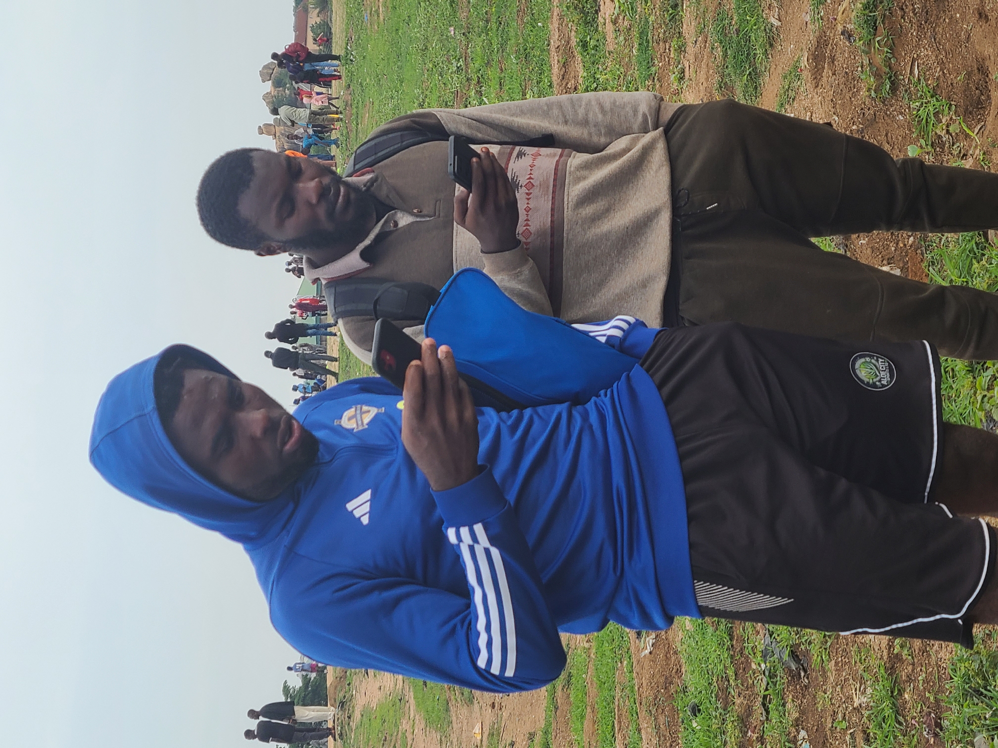 The image shows two men standing outdoors, likely in a field or grassy area. Both men are looking at their phones. The man in the foreground is wearing a blue Adidas hooded sweatshirt and black shorts with white trim and a logo on the right leg. The man in the background is wearing a beige long-sleeved shirt with a patterned chest panel and dark pants. There are other people visible in the background, further suggesting the men are at a gathering or event. The image was taken in Unknown, Unknown.