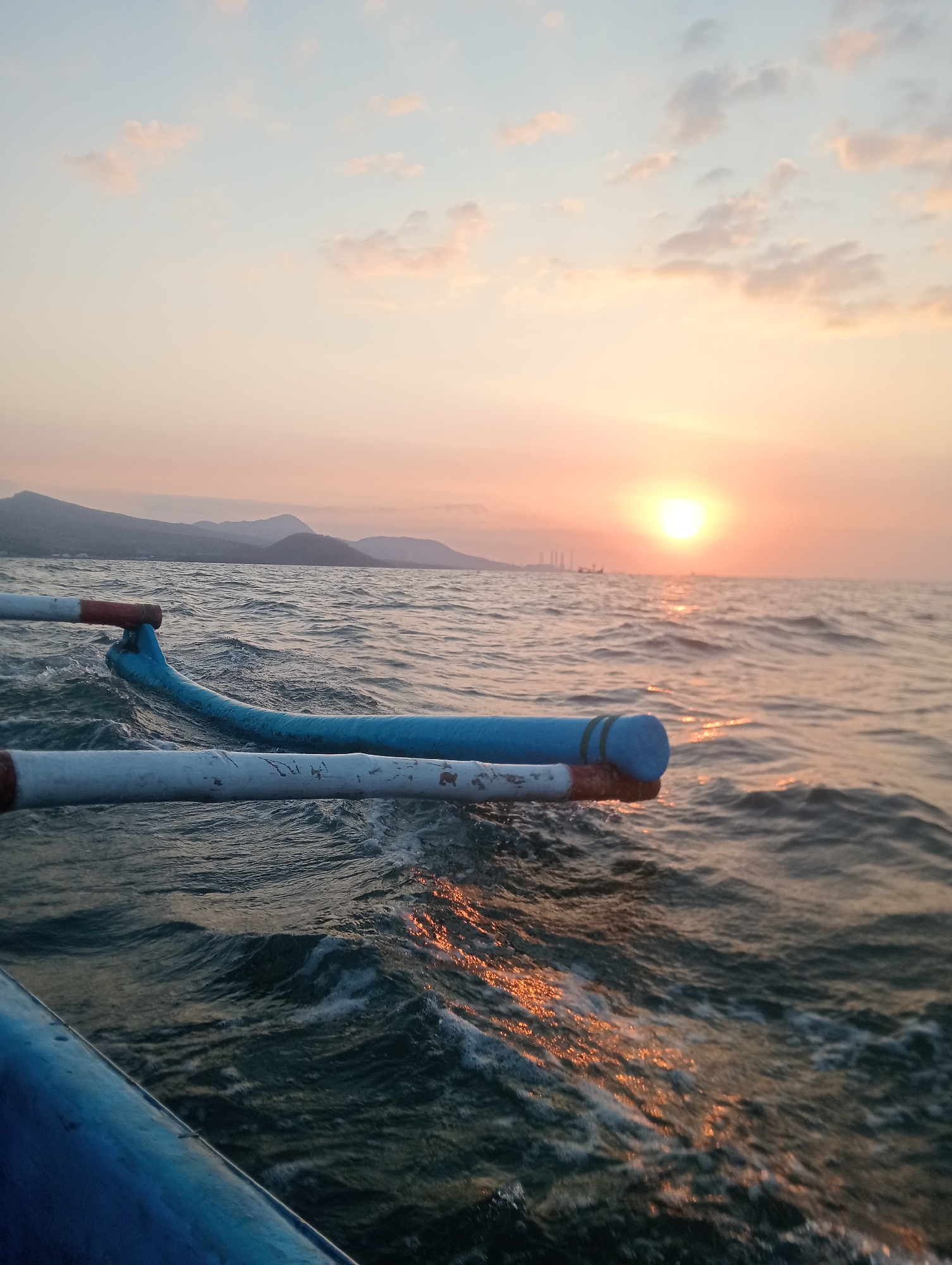 A low-angle, close-up shot captures two outrigger canoe paddles, painted blue and white with red accents, resting on the edge of a boat. The boat is moving through choppy, dark blue water, generating white foam and spray. The sun is setting on the horizon, casting a warm orange glow across the sky and reflecting on the water's surface. In the distance, a hazy silhouette of mountains lines the coast, and a few industrial structures with tall smokestacks are visible. The scene appears to be taking place at sea near the city of Pesisir, Indonesia, likely during a sunset voyage.