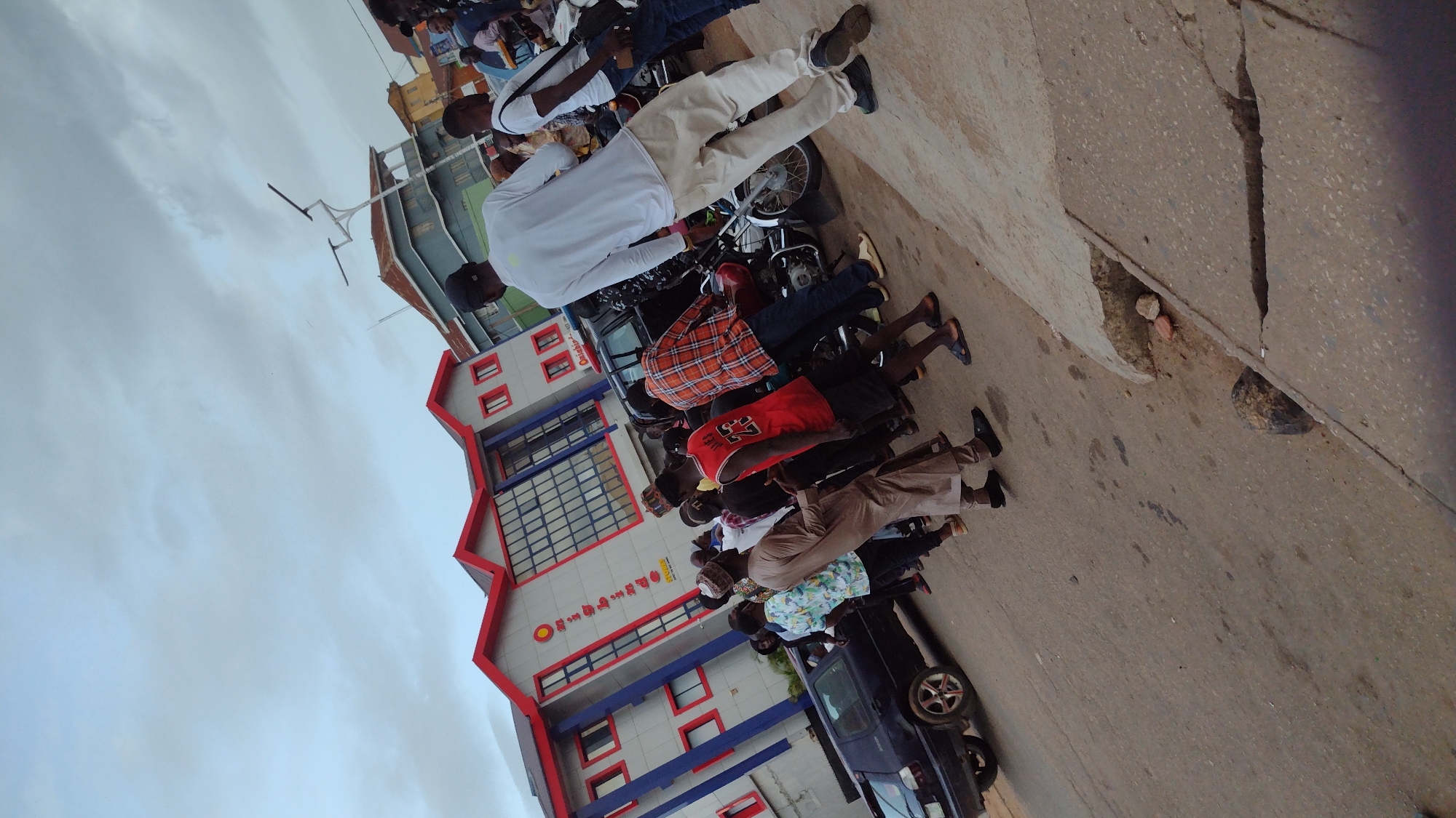 The image depicts a street scene in Jos, Nigeria, with a modern, multi-story building featuring a red and grey facade. A group of people, predominantly young men, are gathered on the street in front of the building. Some are standing, while others are leaning on or near motorcycles. The ground is paved, and a cracked concrete sidewalk is visible in the foreground. The sky is overcast, suggesting cloudy weather. The building's signage, though partially obscured, includes the word "Onigbinde" and "NGCS". The overall atmosphere appears to be one of casual assembly or interaction.