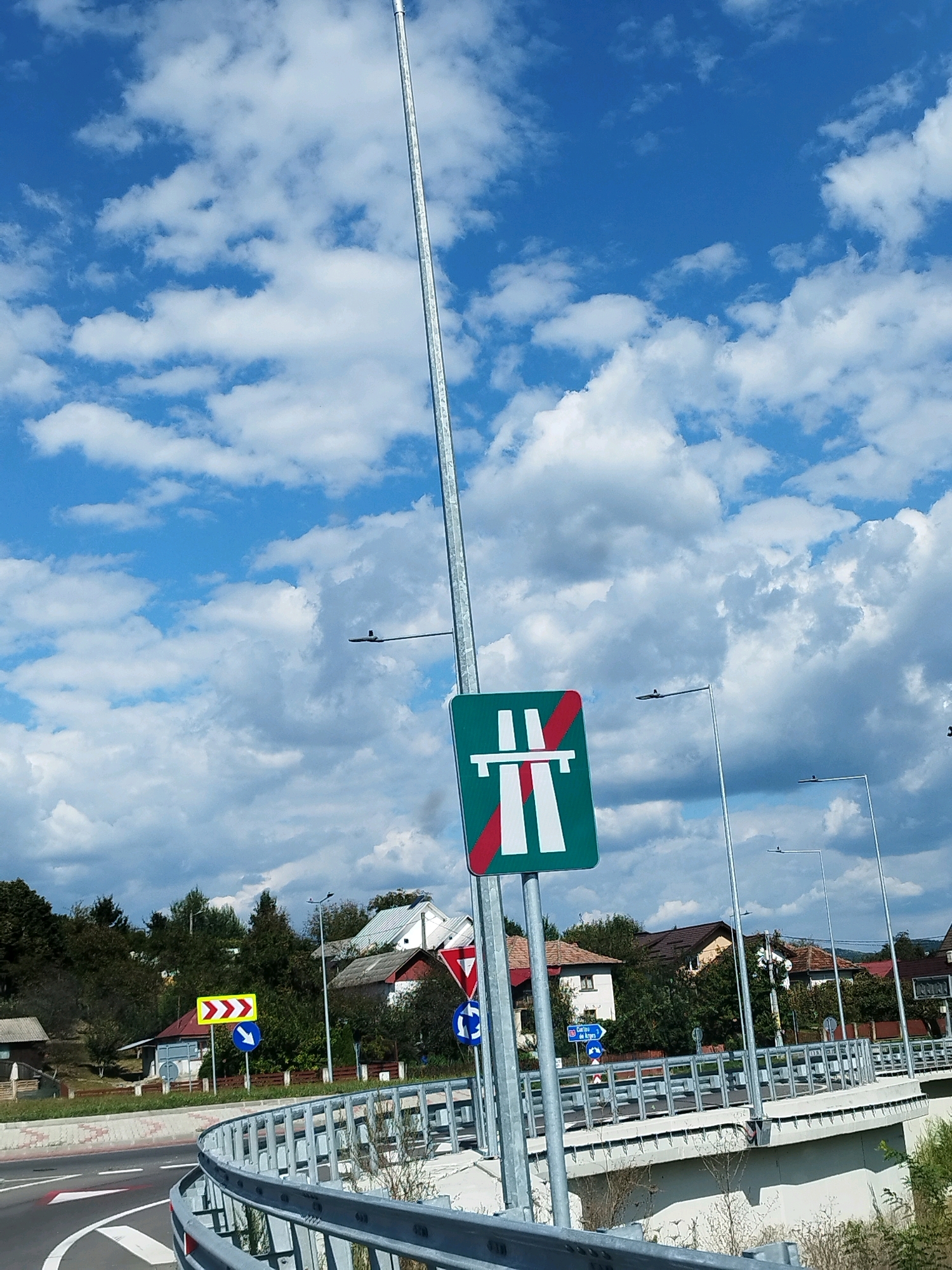 The image shows a highway exit sign in Curtea de Argeș, Romania. The sign, which is green with white markings, indicates the end of the highway. Several other traffic signs are visible, including a directional arrow and a yield sign. The scene is set against a backdrop of blue sky with white clouds, and some houses and trees can be seen in the distance. The highway itself curves to the left, with a metal guardrail along the edge.