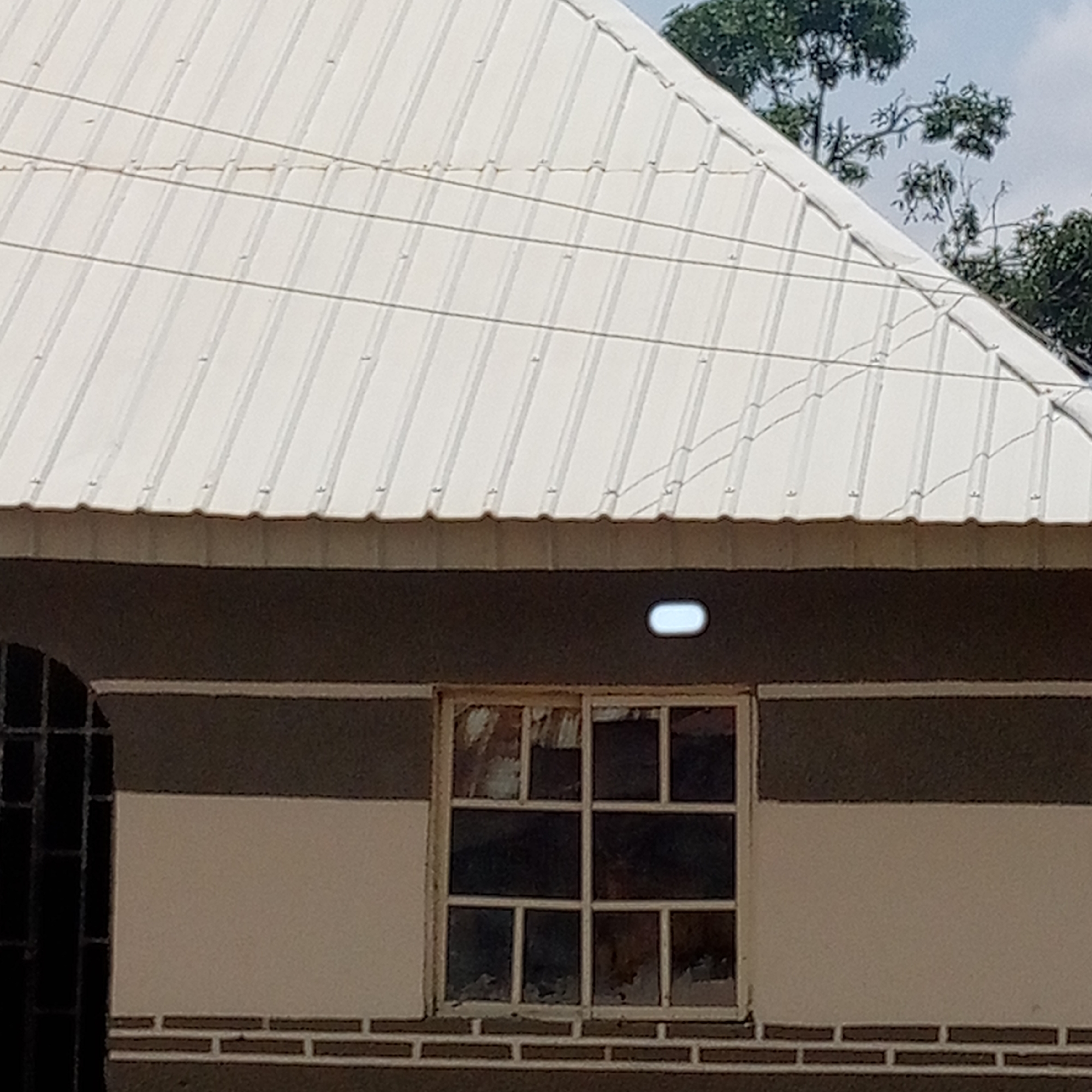 A partial view of a building is presented in the image. The building has a white corrugated metal roof. Below the roof, the exterior wall is painted in a two-tone pattern, with a dark band above a light band. A multi-pane window is visible on the light band of the wall. A security grill is partially seen on the left side of the frame. Trees are visible in the background. Location: Unknown, Unknown.