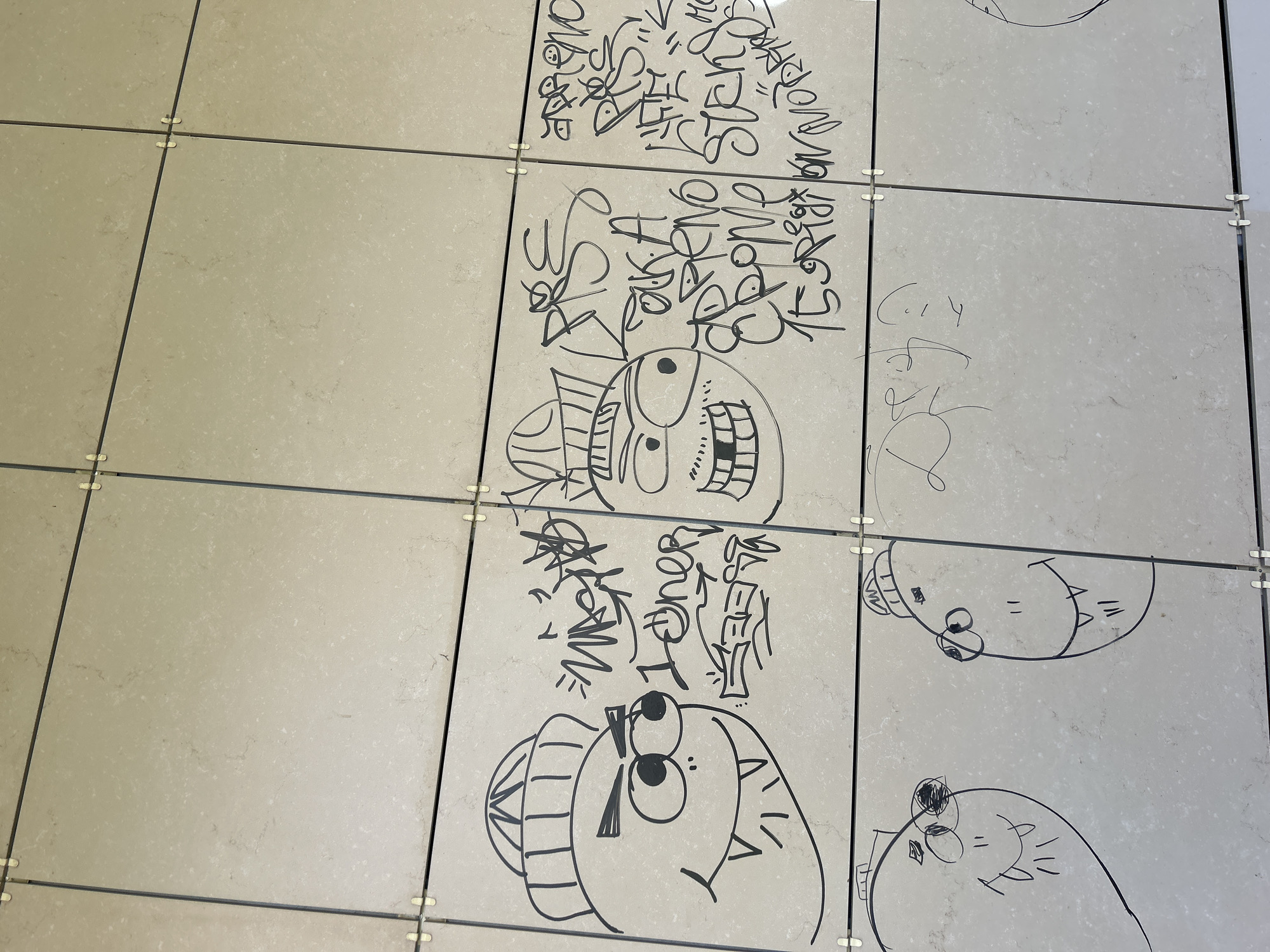 The image depicts a section of an exterior tiled wall, likely part of a building in the city of Chaikovsky, Russia. The wall is composed of rectangular, light-colored tiles with dark grout lines. Several graffiti tags and cartoonish drawings in black marker adorn the tiles.
On the upper left, a cartoon character with a beanie hat, angry eyebrows, large eyes, and sharp teeth is drawn. To its right, a series of graffiti tags are visible, including "MAGNA" and "1oner".
In the upper right section, another cartoon character with a hat, wide eyes, and a large mouth filled with teeth is depicted. Next to it, more graffiti tags are present, with some legible words such as "PRS", "AKA", "EPHEMO", "STENS", and possibly "MORANO".
In the lower left, two more cartoon figures are drawn, both with simple oval shapes for heads, large eyes, and sharp teeth, one with a tongue sticking out. In the lower right section, a section of the wall shows faint, illegible scribbled markings. The overall scene suggests an urban environment with instances of street art and vandalism.