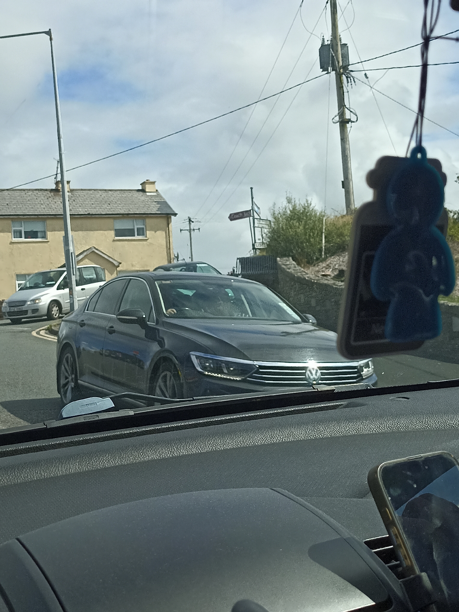 The image shows a street scene, likely in Derrybeg, Ireland, with several cars and buildings visible. In the foreground, the interior of a car is visible, including a dashboard and a phone mount with a phone. Outside the car, a dark-colored sedan is prominent, with its front grill and headlights visible. To the left, a silver car is parked or moving, and further back, a light-yellow building with a tiled roof and windows is present. A tall pole with electrical equipment and wires stands to the right of the frame. The sky is cloudy with patches of blue. The presence of street signs and the architectural style of the buildings suggest an Irish setting. The overall scene depicts a typical roadside environment on a cloudy day.