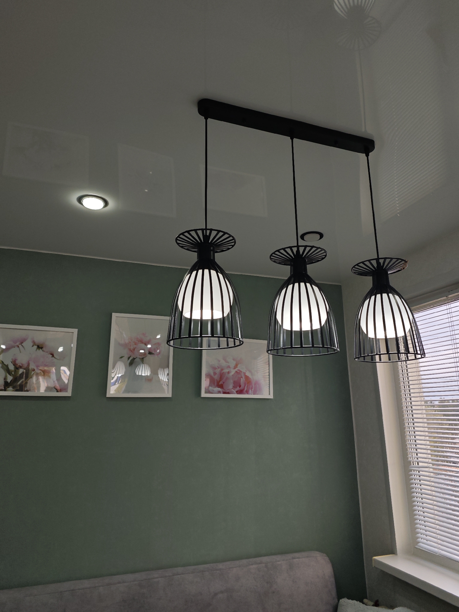 This image captures the interior of a modern room, likely in a home in Фоки, Russia. The primary focus is on the ceiling and a sage green wall, featuring contemporary lighting fixtures and framed art.
Three black pendant lights with conical, slatted metal cages and white inner diffusers hang prominently from a long black bar attached to the white ceiling. Their bright, warm light illuminates the space. To the left, a single circular recessed spotlight is also active on the ceiling. The white ceiling has a glossy finish, reflecting the light fixtures and subtle architectural details.
The wall, painted a distinctive muted sage green, serves as a backdrop for three framed pictures. These artworks appear to depict soft pink floral arrangements, possibly peonies, in a light, airy style. Reflections are visible on the glass of these picture frames, showing parts of the room or the overhead lights.
In the lower left, a portion of a grey upholstered sofa or couch is visible, suggesting a comfortable living or dining area. To the right, a window with closed white horizontal blinds allows some ambient light into the room, although the outdoor view is obscured. The presence of both natural light from the window and active indoor lighting suggests it might be overcast, evening, or simply a preference for additional illumination. No people are visible, and there is no specific activity or event taking place. No visible text is present in the image.