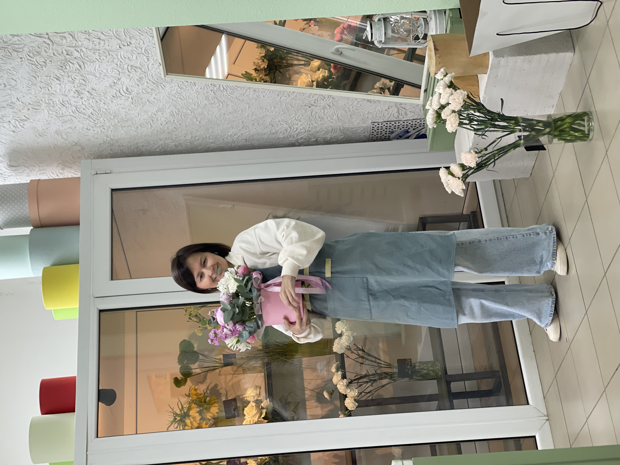 A full-length shot depicts a woman holding a bouquet of flowers. The woman has short, dark hair and wears a white sweater and a light blue denim apron. She holds a pink floral box in front of her with both hands. She smiles.
The shot appears to be indoors, possibly in a flower shop, suggested by the floral arrangements seen through the glass window behind the woman. Various rolls of colored paper are positioned against the wall to the left. A mirror reflecting floral arrangements can be seen in the upper part of the frame. The floor is tiled in a light color. A vase of white carnations is also in view on the right side of the frame. The location is in the city of Unknown, Unknown.