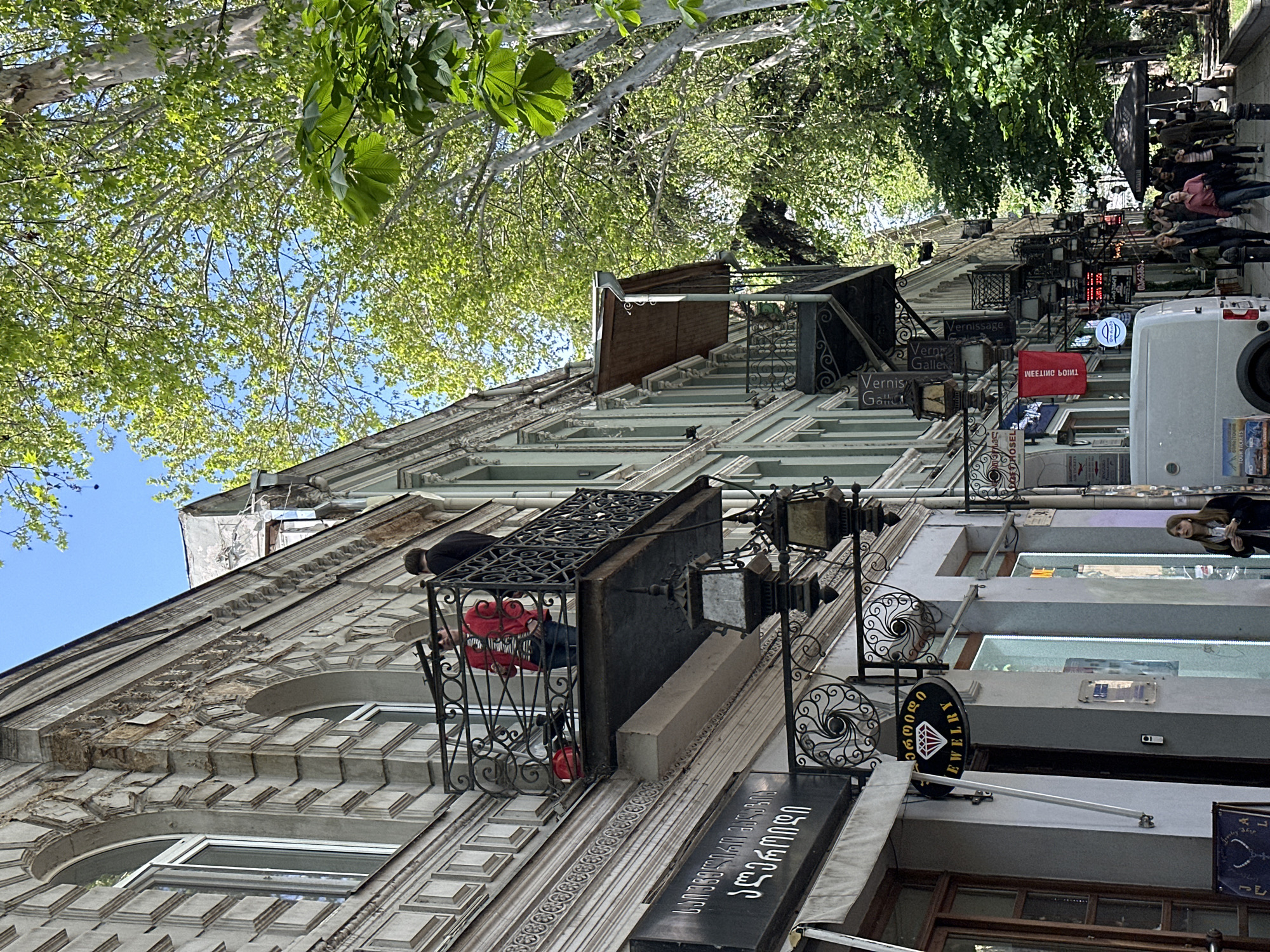 The image shows a street in Tbilisi, Georgia. The perspective is angled upwards towards the buildings lining the street, with trees visible overhead. The buildings are old and ornate, with decorative facades and wrought iron balconies. Several signs are hanging from the buildings, advertising various businesses, including jewelry stores and art galleries (Vernis Galle). The signs are written in both Georgian and English. People are walking along the sidewalk. A white van is parked on the street. A person is visible on a balcony wearing a red jacket.