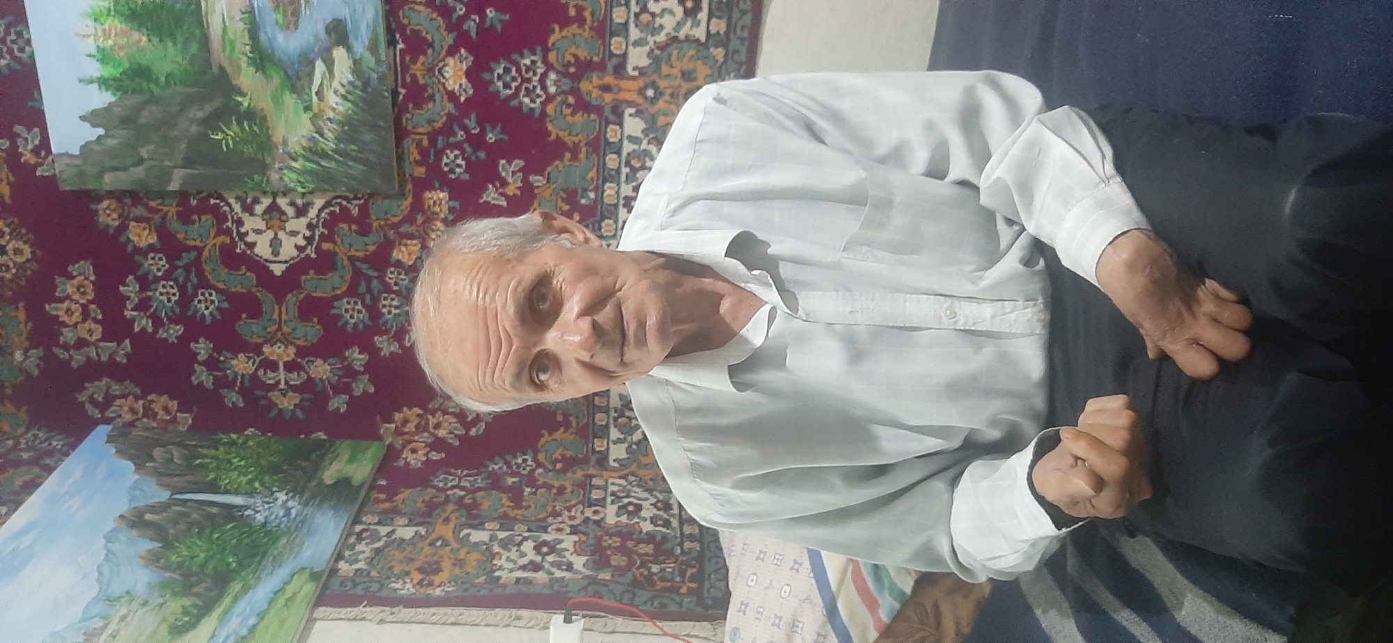 In this image, an elderly Caucasian man is seated indoors, possibly in his home in Yessentukskaya, Russia. He is wearing a light-colored, long-sleeved button-down shirt and dark trousers. The man has gray hair, prominent wrinkles on his forehead and face, and is looking towards the camera with a neutral expression. His hands are resting on his lap, with his left hand clasped over his right.
Behind him, the background features a richly patterned rug with burgundy and intricate designs. To the left of the man, two landscape paintings are visible. The upper painting depicts a forest scene with a stream, while the lower one shows a waterfall cascading down rocks amidst lush greenery under a bright sky. The overall setting suggests a cozy, domestic environment. The lighting appears to be natural, indicating it could be daytime. There is no visible text in the image.