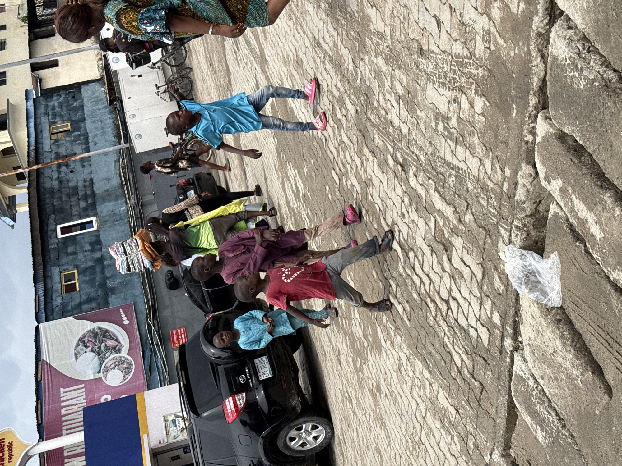 This image shows a street scene in Ado, Nigeria. There are several people, mostly children, standing or walking on a paved area. Some individuals appear to be waiting. A black SUV is parked nearby with people around it. A food banner for "N R Restaurant" and signs for "Chicken Republic" are visible, indicating nearby businesses. The sky is overcast.