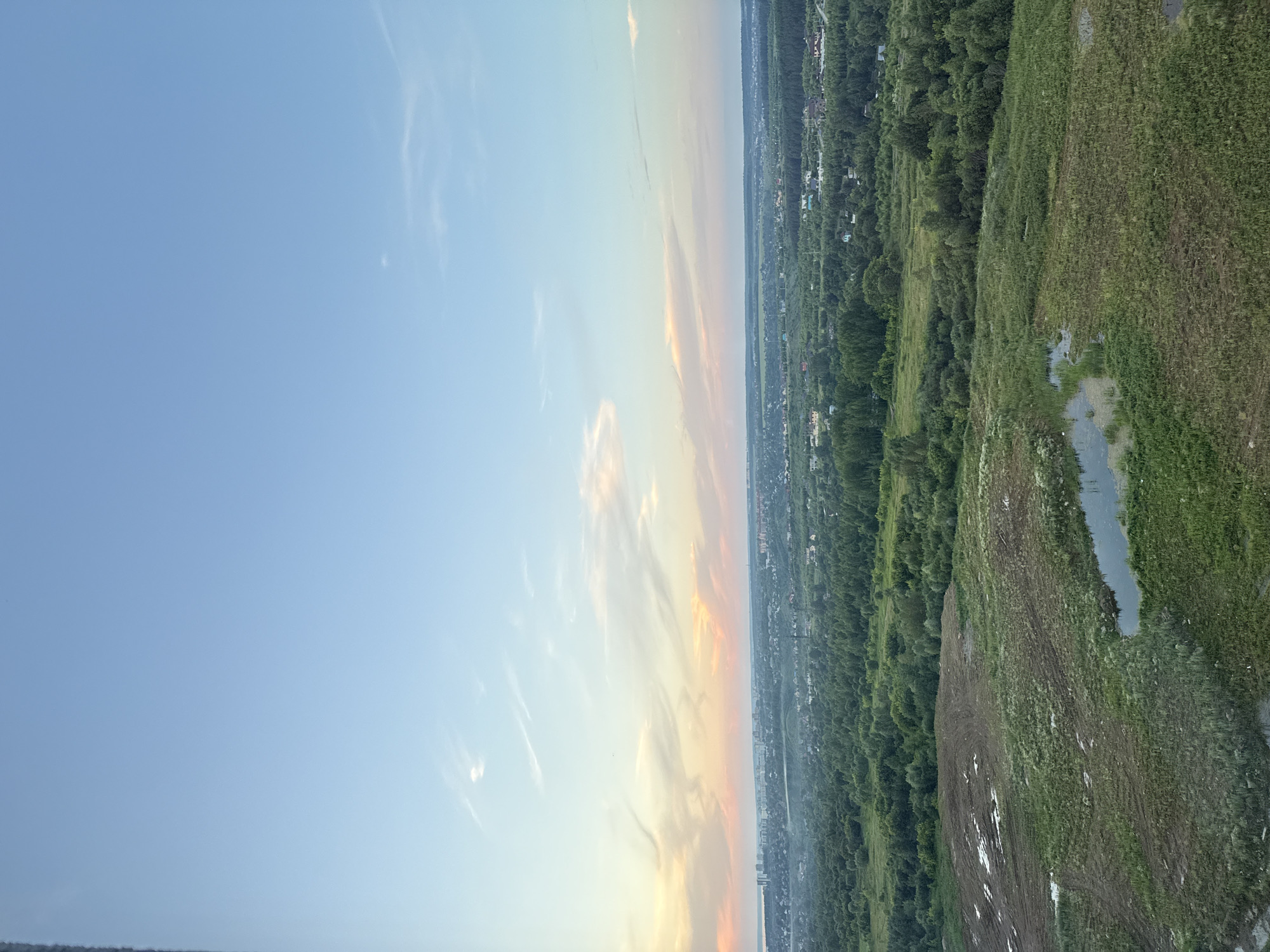 An aerial, wide-angle landscape shot captures a sprawling view of a semi-urban and rural environment under a pale blue sky with wispy clouds tinged with sunset colors. In the foreground, a rough, partially cleared field is dotted with patches of water and muddy earth. Beyond this, a dense, verdant forest stretches across the middle ground, interspersed with scattered houses and buildings, suggesting a suburban or semi-rural settlement. A distant cityscape with taller buildings is visible on the horizon, under a hazy, pale pink and orange sunset sky. The overall impression is one of a peaceful transition from urban development to natural landscape, likely taken from a high vantage point overlooking the outskirts of Perm, Russia, given the landscape and architecture.