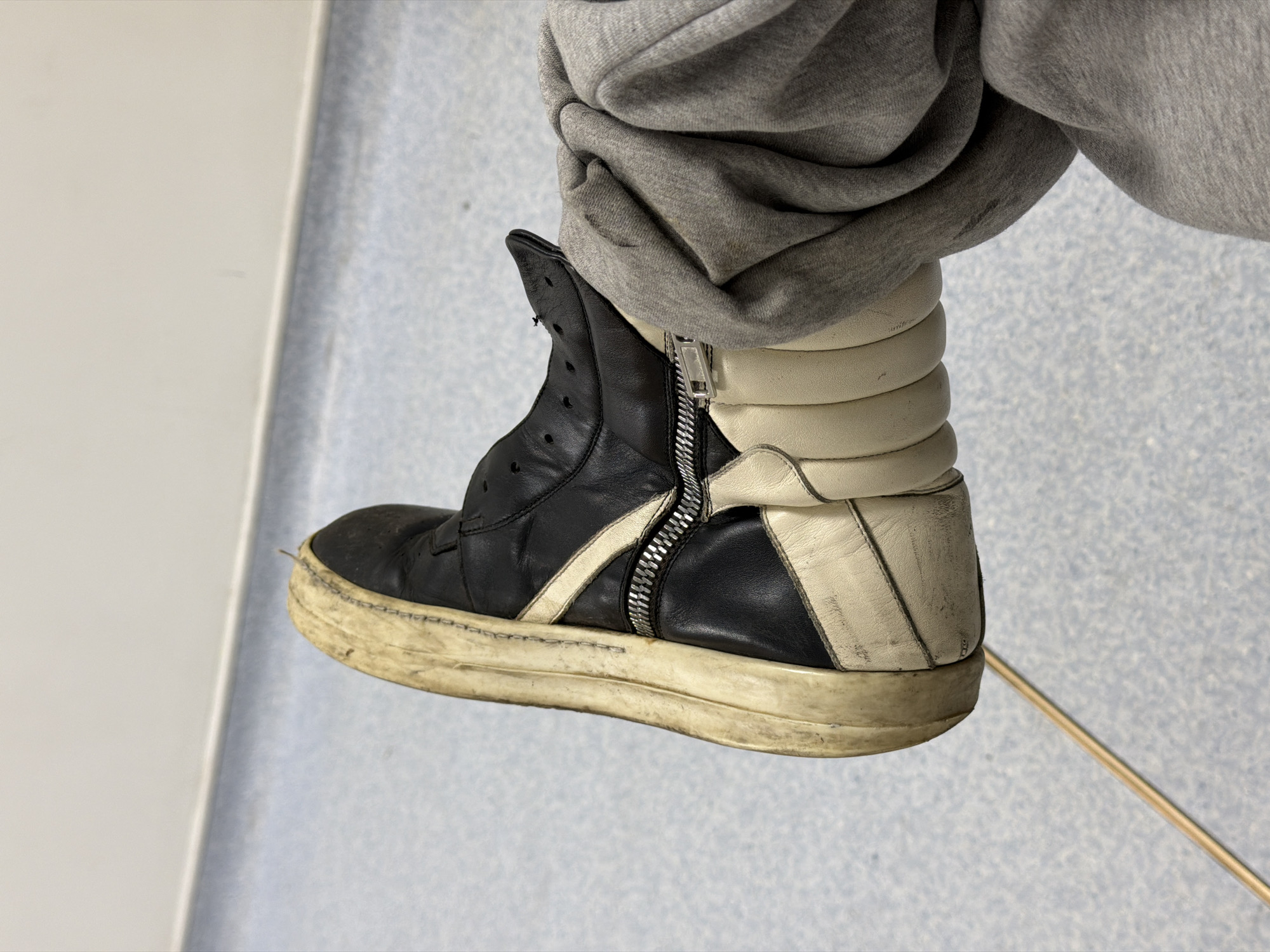 A person wearing a pair of black and white high-top sneakers is standing in an interior space. The shoe is visibly worn and dirty. The person is also wearing gray sweatpants. The background consists of a light blue and white speckled wall and a white wall, suggesting an institutional setting. A metal rod is also visible on the floor. The location of the scene is not discernible from the image. The city of London, United Kingdom is not indicated in this image.
