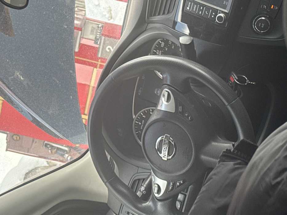 The scene depicts the interior of a vehicle, viewed from a seated position, looking forward through the windshield. A prominent black Nissan steering wheel occupies the central foreground, featuring silver trim, control buttons (e.g., "CRUISE ON/OFF"), and a central Nissan logo. Behind the steering wheel, a portion of the instrument cluster is visible, displaying a digital time of "4:32" and a temperature reading of "33." A set of car keys with a remote fob hangs from the ignition or key slot to the right of the steering column. The leg of an individual, clad in dark trousers or fabric, is visible in the lower left foreground.
Through the windshield, a gas station environment is observed. Multiple red and white striped gas pumps are visible, with one pump clearly displaying the number "2." The ground in the exterior scene shows areas covered with snow or slush. A dark-colored vehicle hood, appearing dusty, is partially visible in the upper central portion of the windshield view. A brown building or structure is partially visible behind the gas pumps on the left. This scene is situated in the city of Apnagyugh, Armenia.