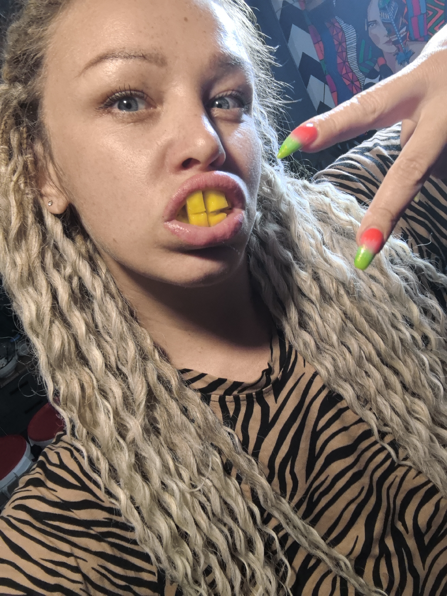 A woman with blonde dreadlocks and blue eyes is holding yellow tic-tac candies in her mouth. She is wearing a black and brown zebra print top. Her fingernails are painted with a red to green ombre effect. The background appears to be a room with patterned artwork on the wall.
