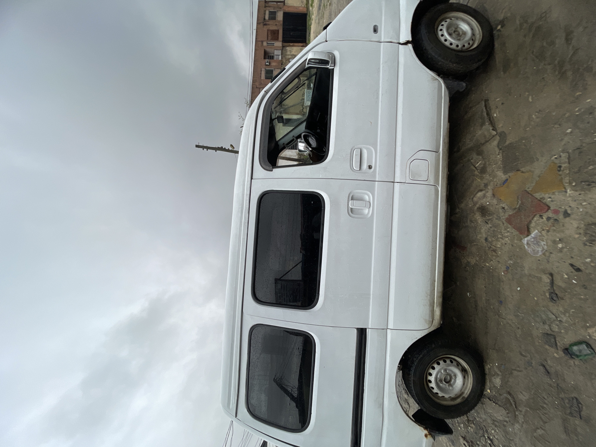 A white van is parked on a paved surface. A building is partially visible in the background. Overcast skies. Location inferred to be Ado, Nigeria.