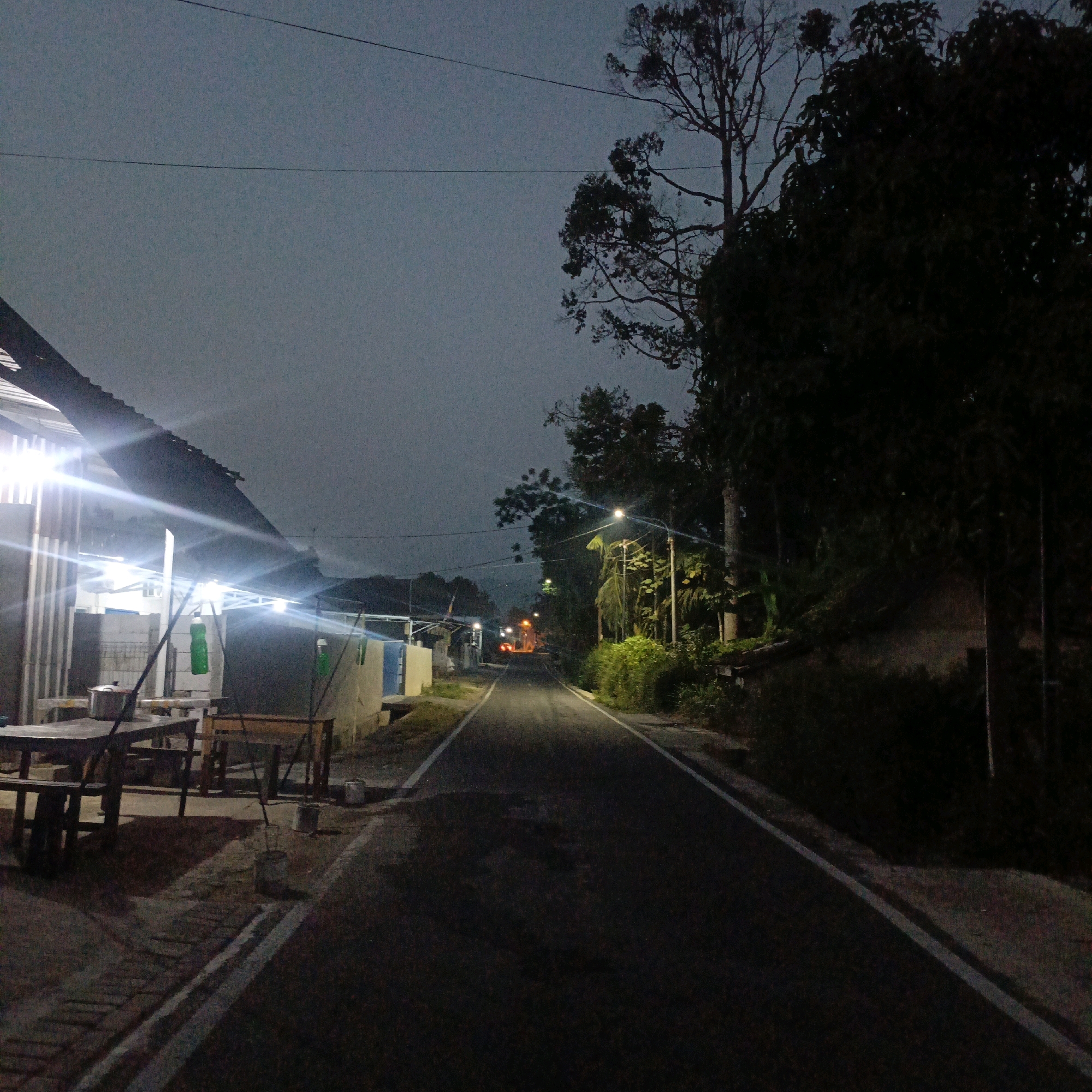 The image depicts a dimly lit asphalt road in Salatiga, Indonesia, at dusk or night. The road is lined with trees and vegetation on the right side, and structures on the left. Streetlights illuminate portions of the road and the surrounding area. On the left, there are buildings with bright artificial lighting, suggesting a commercial area or residences. Tables and chairs are visible outside one of the illuminated structures, possibly an outdoor eatery. Further down the road, distant lights indicate movement or other illuminated structures. The sky is overcast and dark, typical of evening or pre-dawn conditions. No individuals are clearly visible in the scene.
