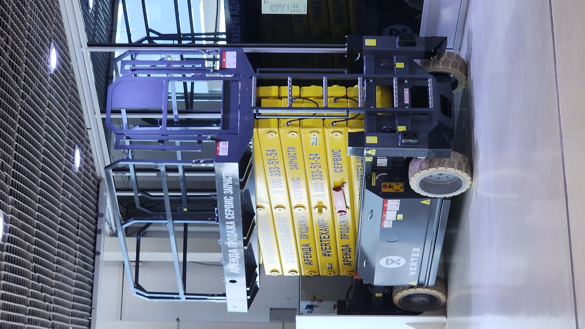 The image shows a grey and yellow scissor lift parked indoors on a polished concrete floor. The scissor lift is in the foreground, with its platform extended upwards. The platform is a dark blue metal structure with railings. The scissor lift has the "VERTEX" logo on its side.
The background shows a modern interior with a tiled ceiling and large windows. The overall environment appears to be an exhibition hall or a showroom. There are no people visible in the image. The location is identified as Moscow, Russia.
