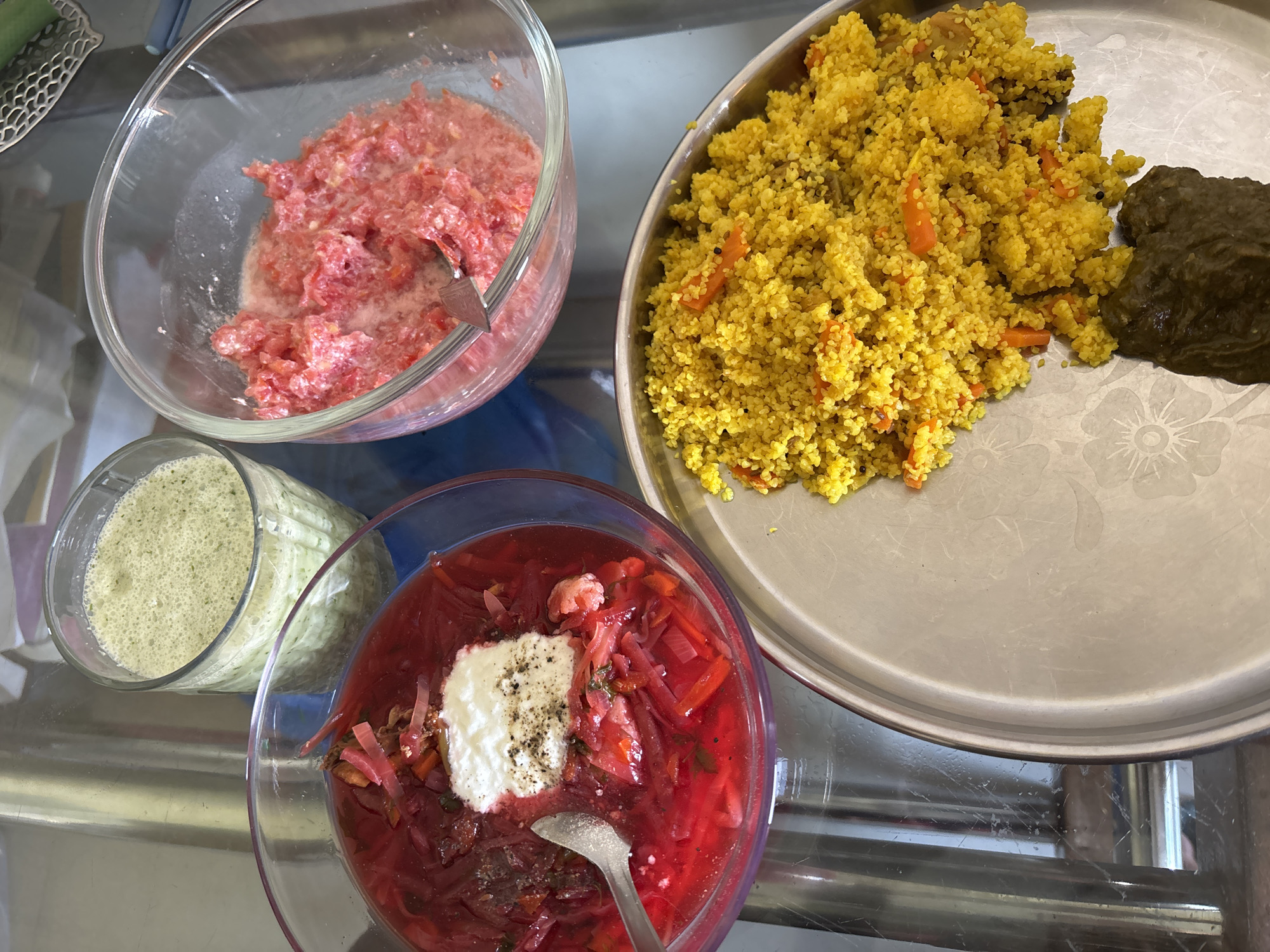 The image shows a variety of food dishes arranged on a reflective surface, likely a glass-topped table. There are four distinct containers:
1. A clear glass bowl filled with a coarsely chopped mixture of red ingredients, possibly tomatoes or beets, suspended in a milky-white liquid. A utensil, likely a spoon, is inside the bowl.
2. A clear glass jar containing a light green, frothy substance, possibly a blended drink or sauce.
3. A clear glass bowl of red soup, likely borscht, with visible shredded beets and topped with a dollop of white cream and black pepper. A spoon is inside the bowl.
4. A round metal tray holding a pile of yellow couscous-like grains with chopped carrots and a dark green, possibly cooked, vegetable item.
The table is situated indoors under what appears to be natural lighting. The location is specified as Ambernath, India, but this context is not discernible within the image alone.