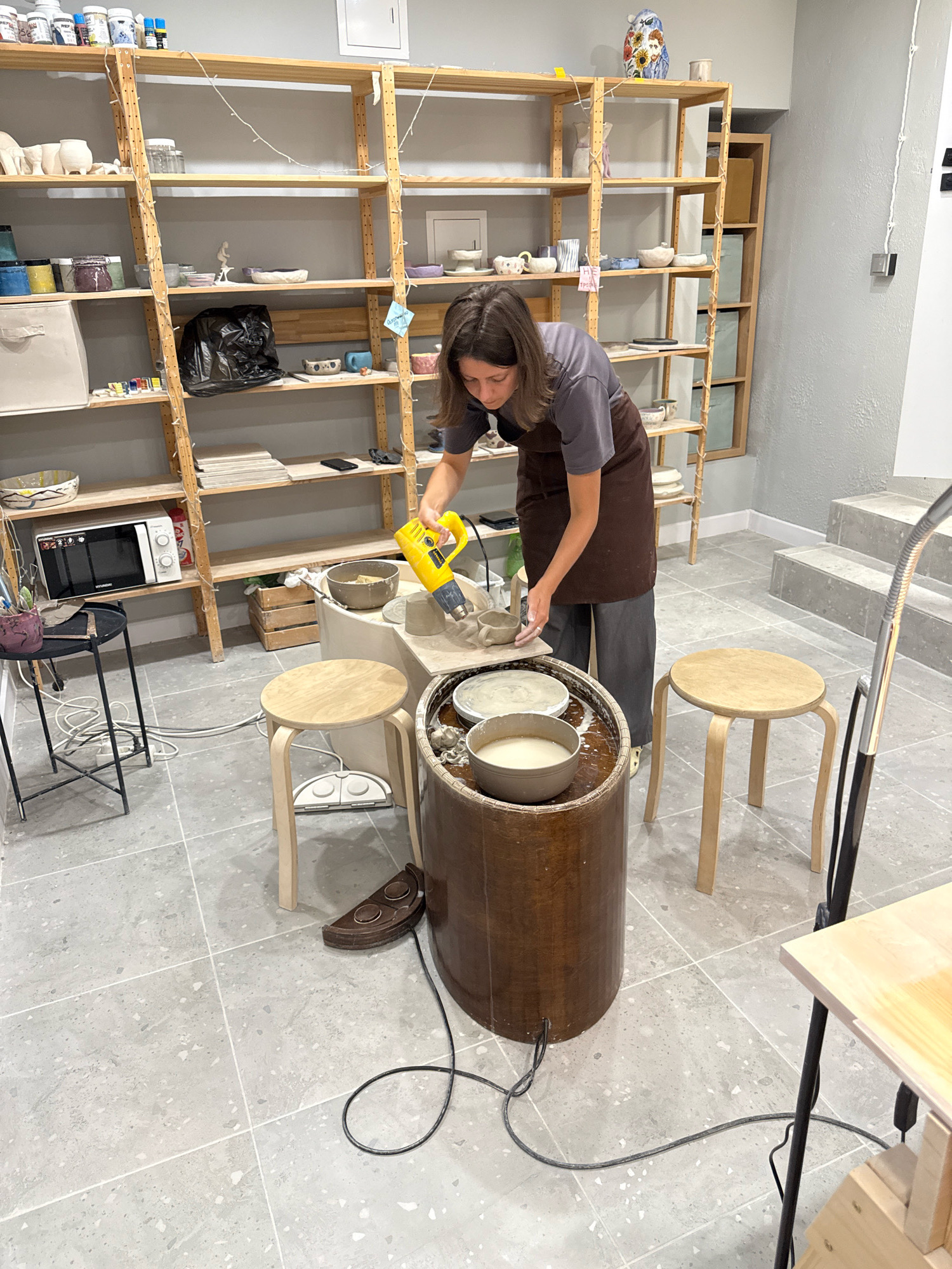 A woman in a brown apron and gray pants uses a yellow heat gun on a pottery wheel in a studio. The studio has shelves filled with pottery supplies and finished pieces. A microwave is on a shelf to the left of the woman. There are three stools and a large wooden cylinder. The floor is made of gray terrazzo tiles. The image was taken in Saint Petersburg, Russia, at the "Art-Pottery" studio.