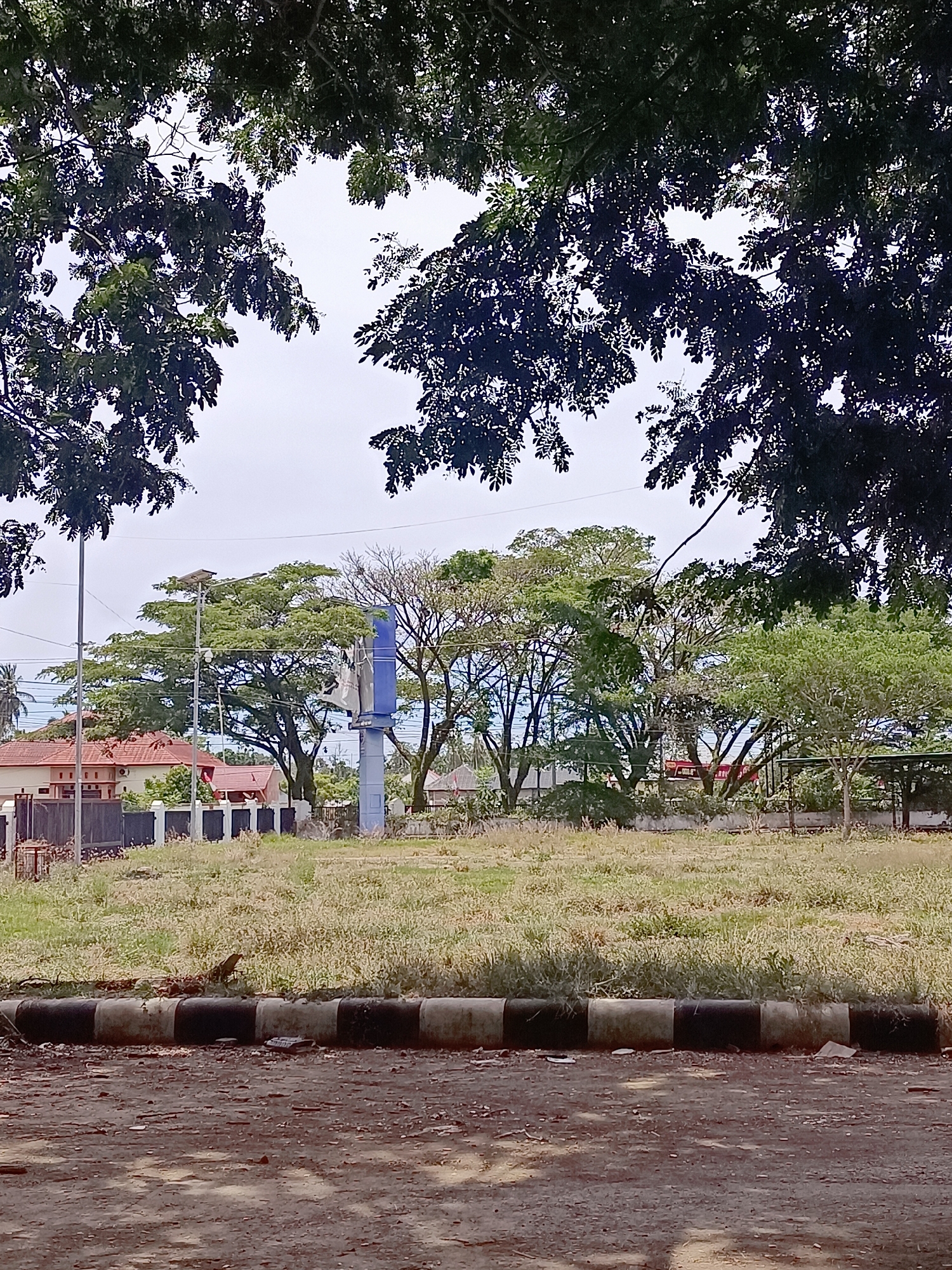 In the city of Meunasah Pupu, Indonesia, the image captures an outdoor scene dominated by lush green trees framing the upper portion. Below the canopy, an open, grassy field is visible with scattered patches of dry vegetation. In the middle ground, a tall blue and white structure, possibly a billboard or signpost, stands against a backdrop of various trees. To the left, a building with red-tiled roofs and a dark fence is partially visible. A solar-powered street light is also present near the building. The foreground features a dirt path with a decorative curb composed of alternating black and white segments. The overall atmosphere suggests a clear day with diffused light, possibly overcast.