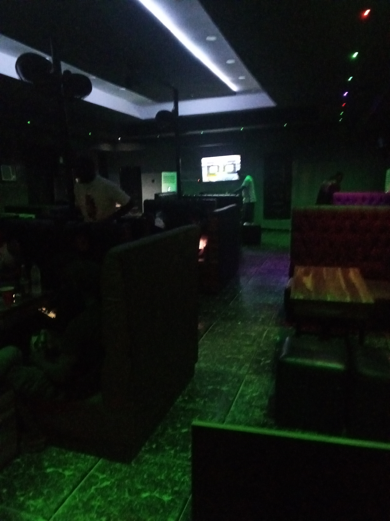 The image depicts the interior of a dimly lit establishment, possibly a lounge or club. The primary illumination comes from green and white LED lighting, casting an eerie glow on the tiled floor and dark furniture. In the background, a large screen displays a muted image, with figures visible in front of it. Several individuals are present, some appearing to be staff, as suggested by their activity behind a counter. The seating arrangements consist of plush, dark-colored booths, with small tables positioned in front of them. The overall ambiance is one of subdued nightlife.