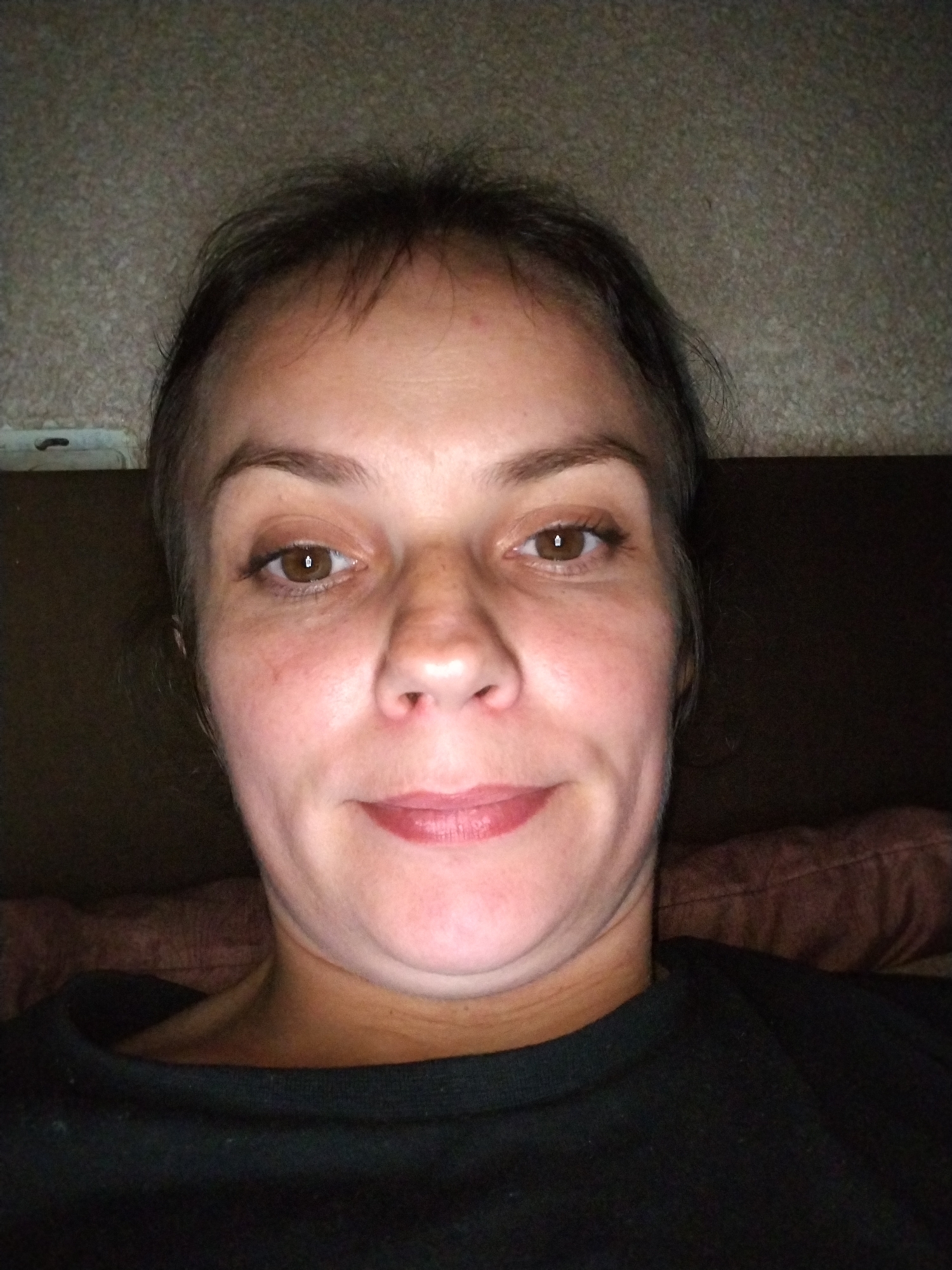 The image is a close-up, overhead, selfie-style photograph of a woman's face and upper chest. She has light brown eyes, fair skin with some visible pores and fine lines, and dark brown hair with lighter brown streaks. Her lips are painted a rosy-pink color. She is wearing a dark-colored, crew-neck shirt. The background appears to be indoors, with a textured wall behind her head and a dark brown, possibly upholstered, surface to her right and behind her shoulders. There is a faint reddish-pink object visible behind her right shoulder, which could be a cushion or blanket. The lighting is dim and appears to be coming from above and slightly in front of her. There is no indication of the location within the city of Molodiia, Ukraine.