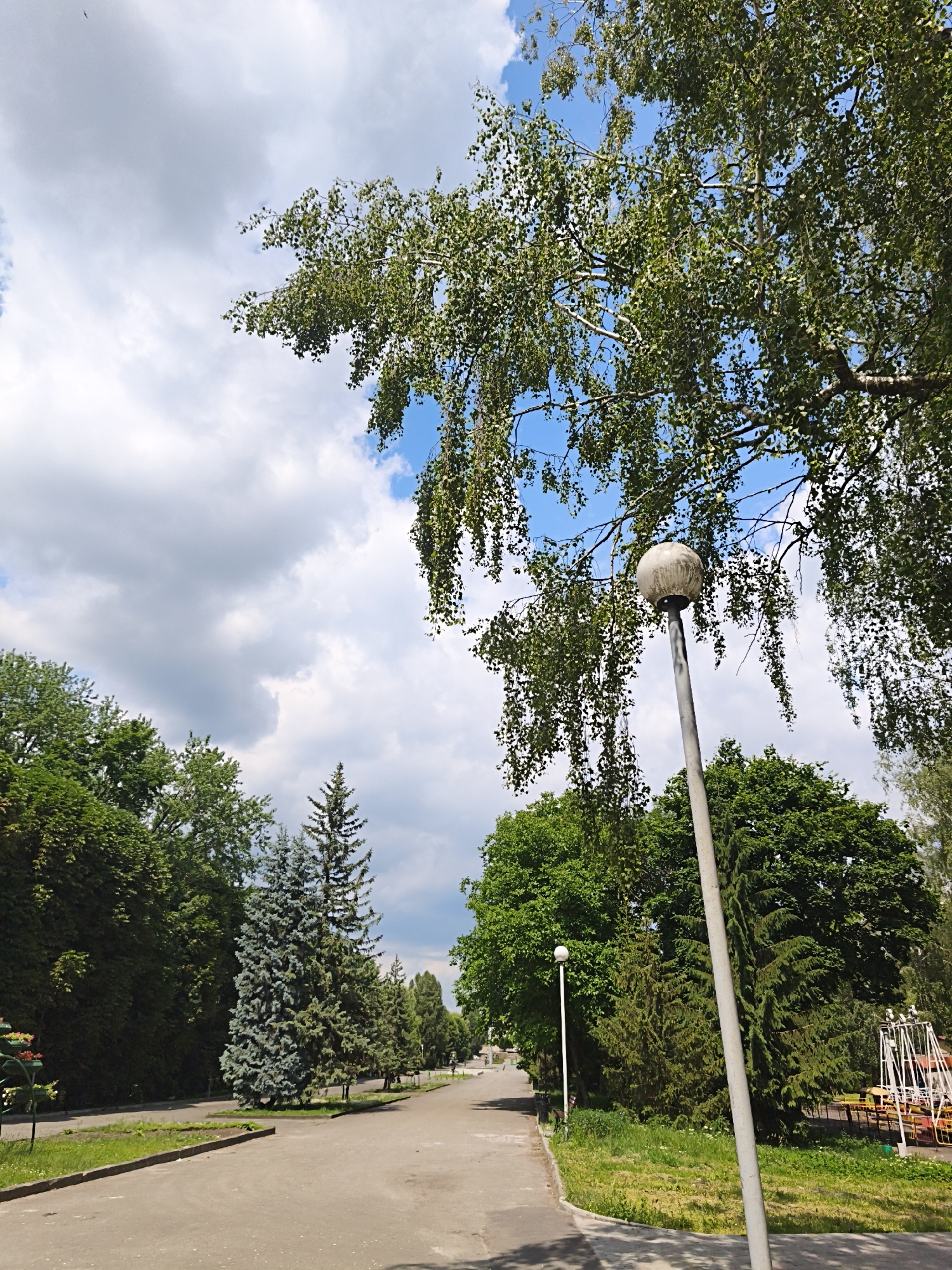 The image depicts a view of a park pathway with a streetlamp in the foreground. The pathway is made of asphalt and is lined with green grass and various trees. In the background, there is a playground with a swing set and tables. The sky is blue with some clouds, and the sun appears to be shining. The scene suggests a peaceful, natural setting within a city. Based on visual cues and common park aesthetics, this could be a typical park scene found in a Ukrainian city like Ternopil.