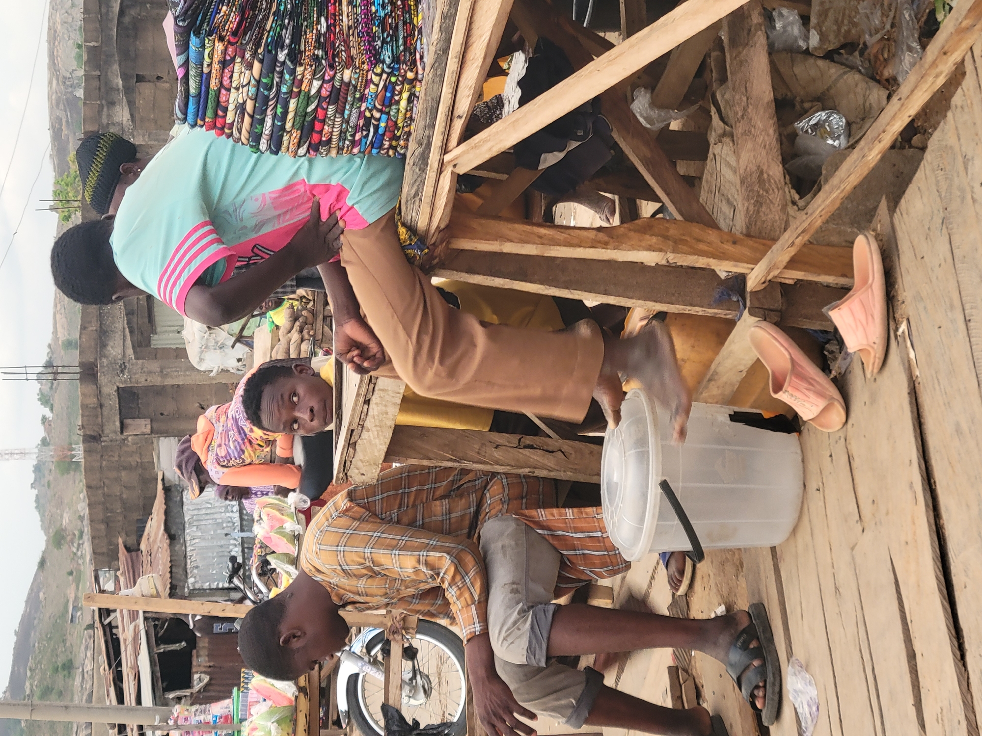 The image shows a street scene in Madalla, Nigeria. In the foreground, two men are on a wooden structure. One man, wearing a plaid shirt and grey pants, sits on the structure, while another man, wearing a light green shirt with pink stripes and tan pants, stands on the structure. There are colorful fabrics stacked on top of the structure. Below the wooden structure is a bicycle, watermelon, and plastic bags. Near the wooden structure are a clear plastic bucket and pink sandals. In the background, buildings and hills can be seen.