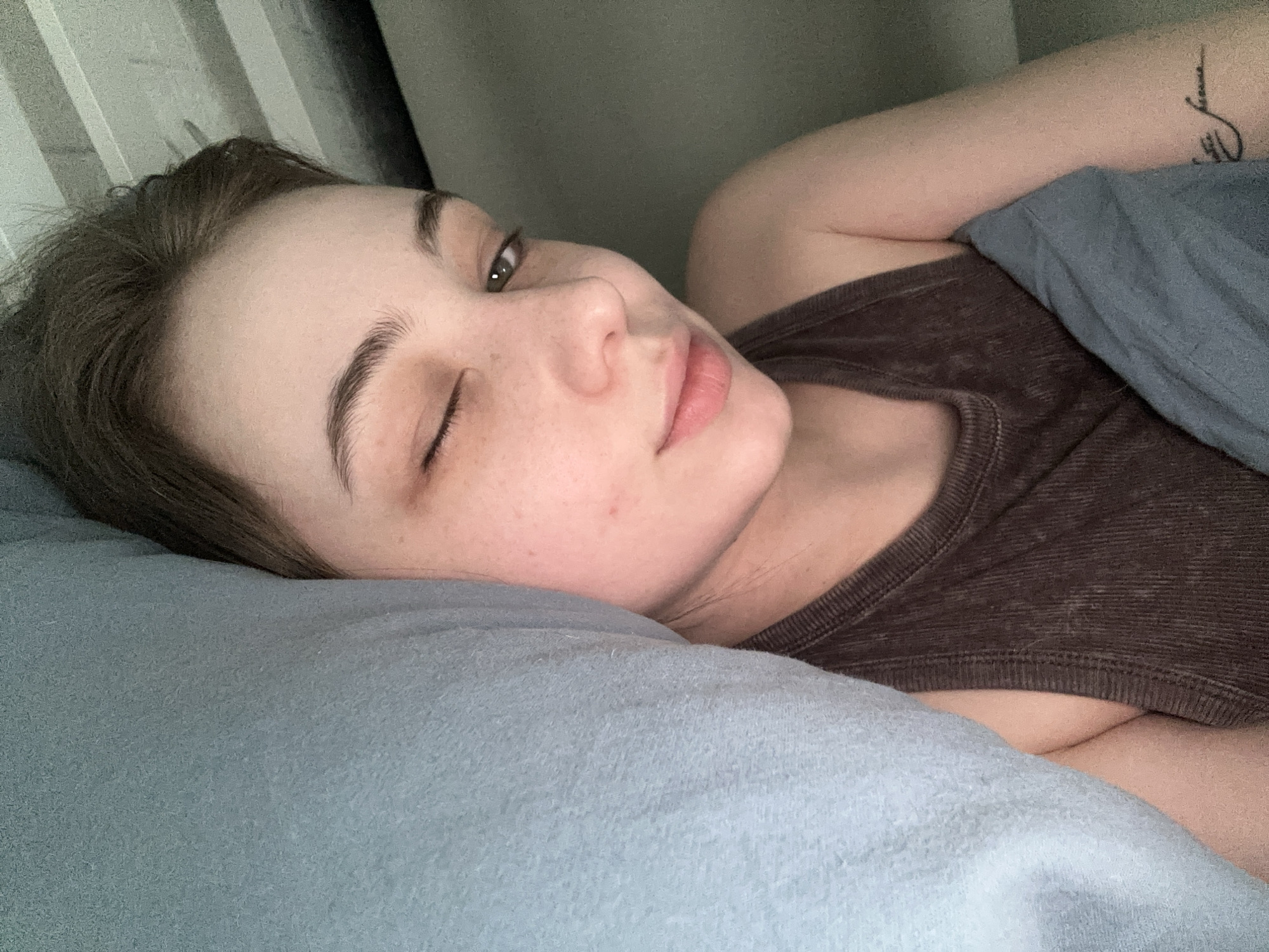 A woman is lying on a blue pillow with a blue blanket draped over her shoulder. She is wearing a brown tank top. One eye is open while the other is closed. There is a tattoo visible on her arm. The background shows a striped wall. The location is not discernible from the provided image.