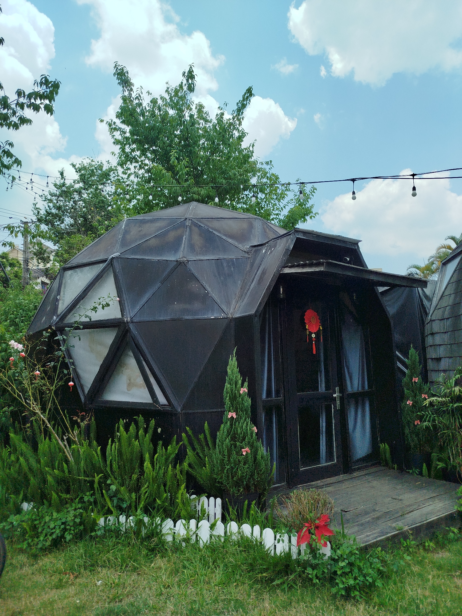 The image shows a geodesic dome structure in what appears to be a garden setting. The structure is primarily black with triangular or trapezoidal sections composing the dome. The dome has windows and an entrance area with a door. A red Chinese New Year decoration hangs on the door. Vegetation, including ferns, small trees, and flowering plants, surrounds the base of the dome, enclosed by a small white picket fence. A wooden platform leads to the entrance. A string of lights is visible overhead. The sky is blue with scattered clouds. The location is likely Da Lat, Vietnam, based on the architectural style and vegetation commonly found in that region.