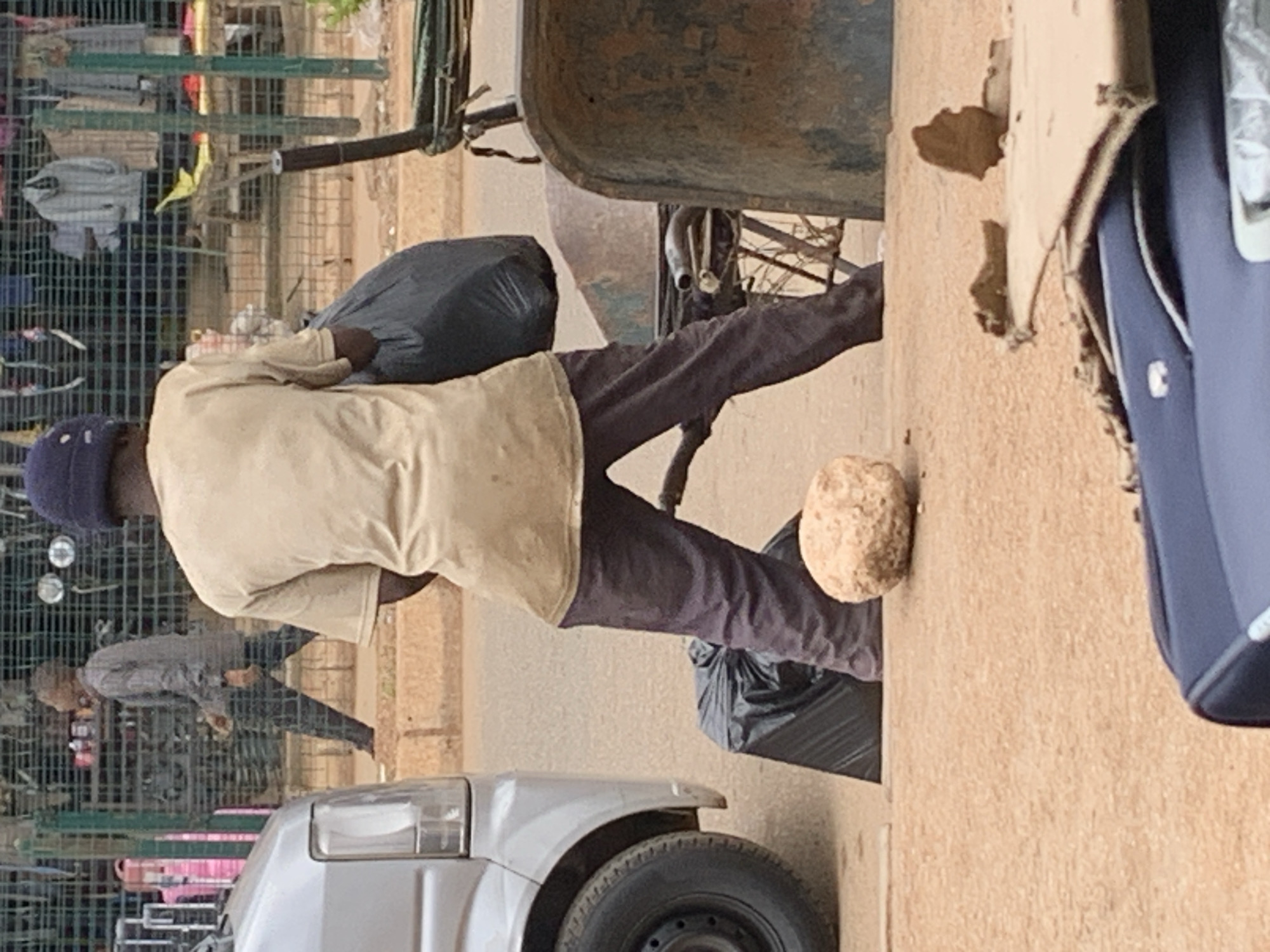 The image shows a person wearing a beige shirt, dark pants, and a blue head covering. The person is carrying a large black bag over their shoulder. The scene appears to be outdoors, with a fence in the background containing various items, possibly clothing for sale. A cart or wheelbarrow with a rusty container is visible behind the person. A light-colored, textured wall is to the right of the person, and part of a blue bag or backpack is also visible. On the ground, next to the person's legs, is a large, round, light-brown object that resembles a rock or a large lump of earth. In the foreground, a portion of a silver car is visible, including its headlight and tire. The overall impression is that of a market or street scene. The lighting suggests it could be daytime, possibly late morning or afternoon, with no obvious indicators of weather conditions. There is no visible text in the image. The image is taken from a slightly elevated perspective and is oriented vertically.
