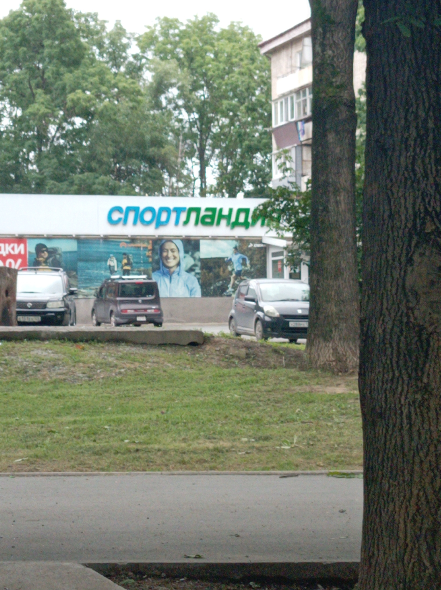 The image captures a street scene in Artyom, Russia, featuring a storefront with prominent Cyrillic signage reading "СПОРТЛАНДИЯ" (Sportlandia). The storefront is adorned with a mural depicting various outdoor activities, including individuals hiking and running. In front of the store, three cars are visible: a dark SUV on the left, a compact purple vehicle in the center, and a dark hatchback on the right. The cars are parked on a paved surface adjacent to a grassy embankment. Trees are a significant element in the foreground and background, with two large tree trunks dominating the right side of the frame. A multi-story building, likely residential or commercial, is partially visible behind the storefront and trees. The overall atmosphere suggests an overcast day, with muted lighting.