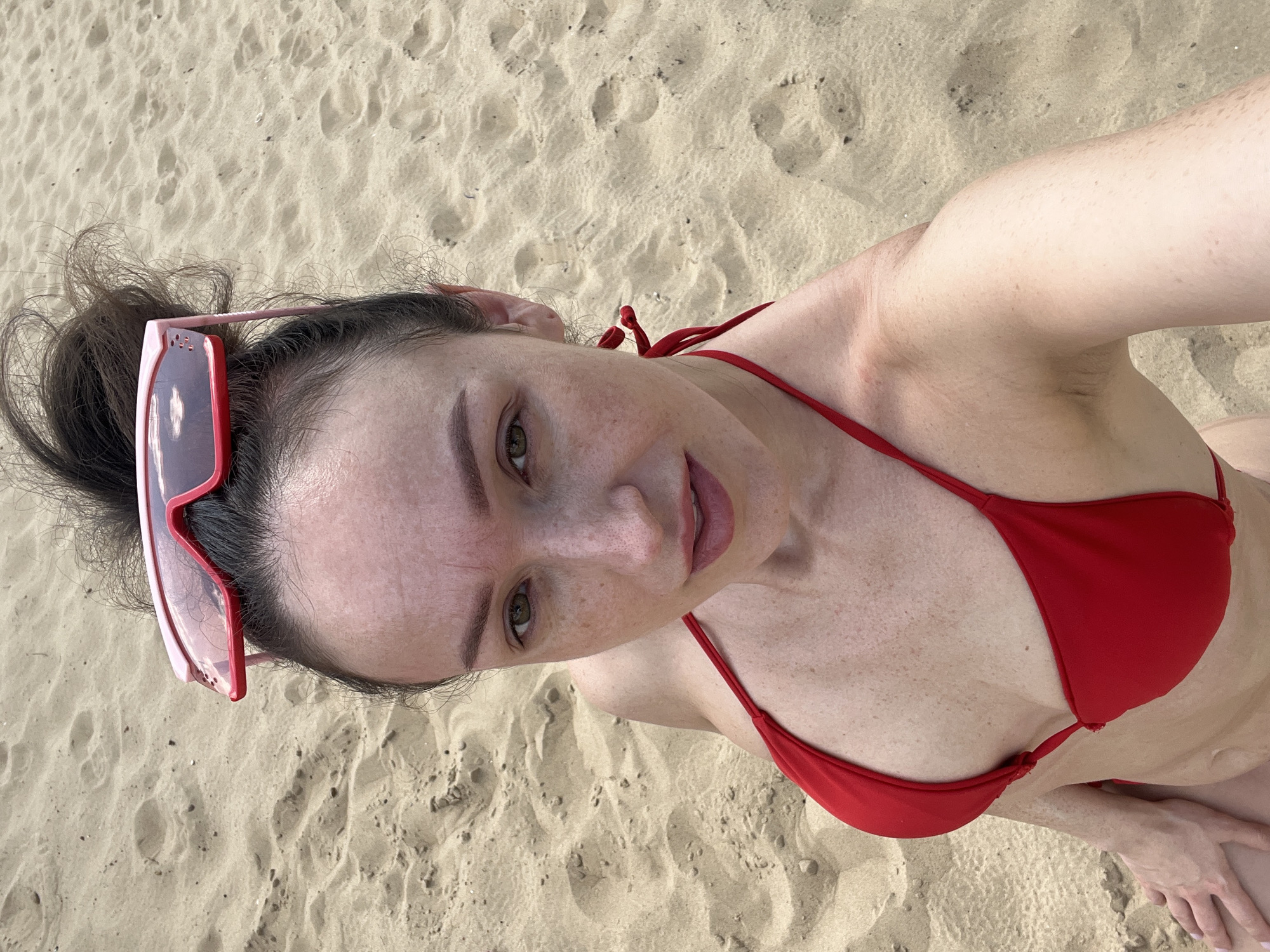 A woman is posing for a selfie on a sandy beach. She has her hair in a messy bun and is wearing red sunglasses on top of her head. She is also wearing a red bikini. The background is the sandy beach. The image was taken in Kazan, Russia.