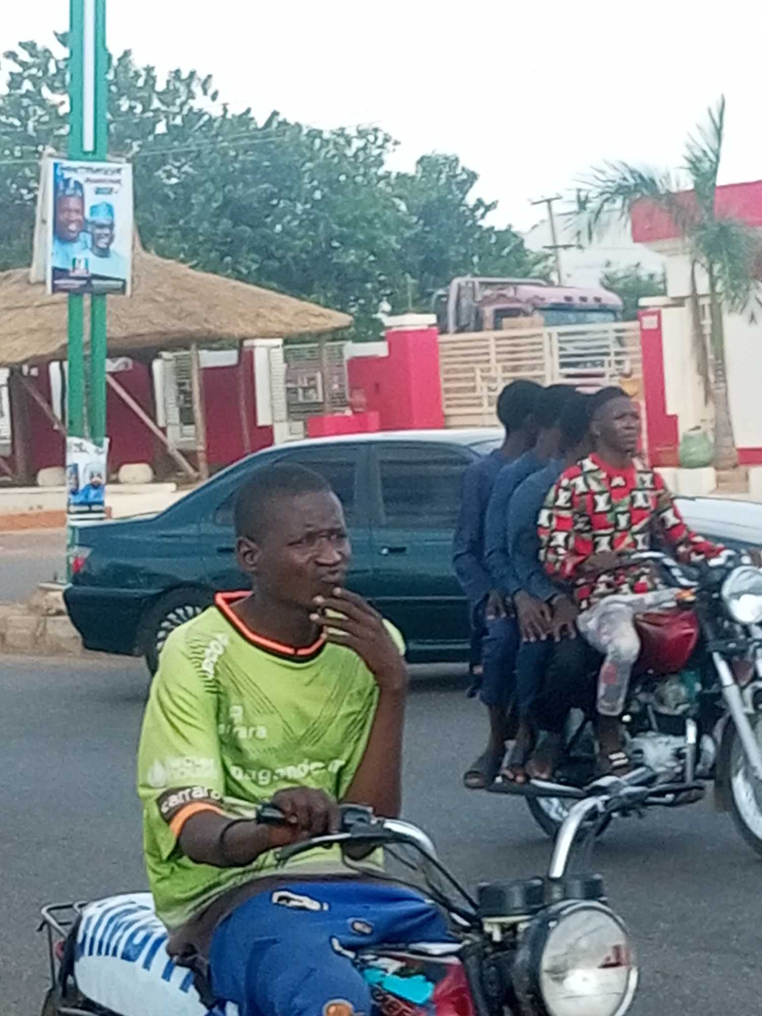 The image depicts a street scene in Garko, Nigeria, with people and vehicles. In the foreground, a young man wearing a green t-shirt is riding a motorcycle. He has his hand resting on his chin, with a thoughtful expression. The motorcycle has a blue and white seat cover.
Behind the first motorcycle, another motorcycle carries four people. The rider wears a colorful, patterned shirt and light-colored pants. Three passengers are seated behind him, all wearing blue shirts. The person at the very back appears to be a young boy.
A dark green sedan is parked on the side of the road. In the background, there are buildings with red accents and thatched roofs, suggesting a marketplace or commercial area. A utility pole with wires is also visible. Further back, a large truck is parked. A poster is attached to a green pole near the thatched-roof structures. The poster features two men in traditional attire, possibly for a political campaign, with some text in a language that appears to be Hausa.
The lighting suggests it is daytime, likely late afternoon, with overcast skies. The overall atmosphere seems to be one of everyday life and transportation in a Nigerian town.