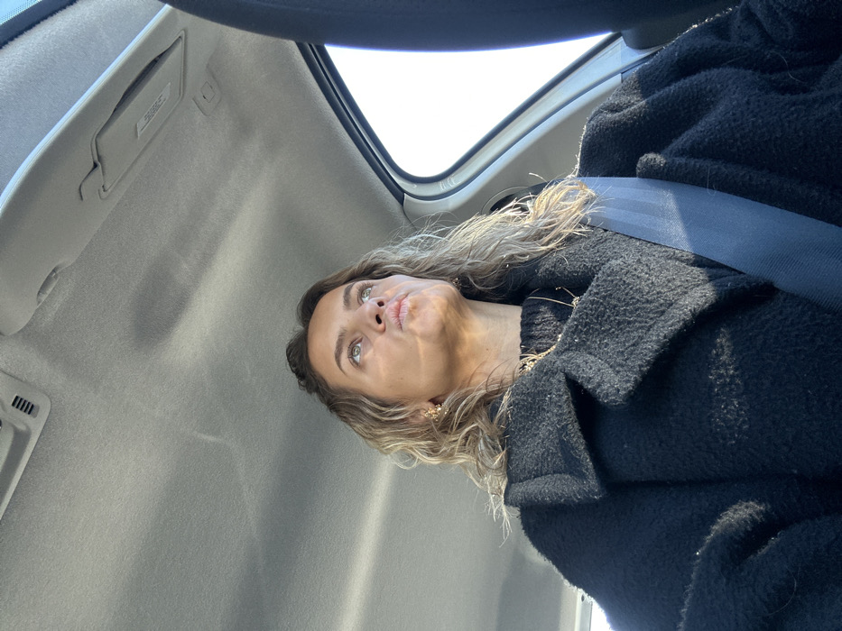 The image depicts a female individual seated within a vehicle, identified as being in the city of Saint-Jean-Bonnefonds, France. The woman has wavy, light brown hair and a light complexion. She is wearing a black, textured overcoat with a collar, over what appears to be a black knitted garment. A delicate gold-colored necklace featuring small, dried, plant-like elements is visible, along with a gold-colored earring on her visible right ear. A blue seatbelt diagonally crosses her torso from left to right. Her head is tilted slightly upwards, with her eyes directed towards the upper right of the frame, and her lips are pursed into a distinct 'fish face' expression. The interior of the vehicle shows a gray headliner with an integrated console element. Strong natural light originates from the right side of the frame, illuminating the woman's right profile and the adjacent vehicle window, which displays a clear, bright sky. No other individuals or external scenery are identifiable within the image.