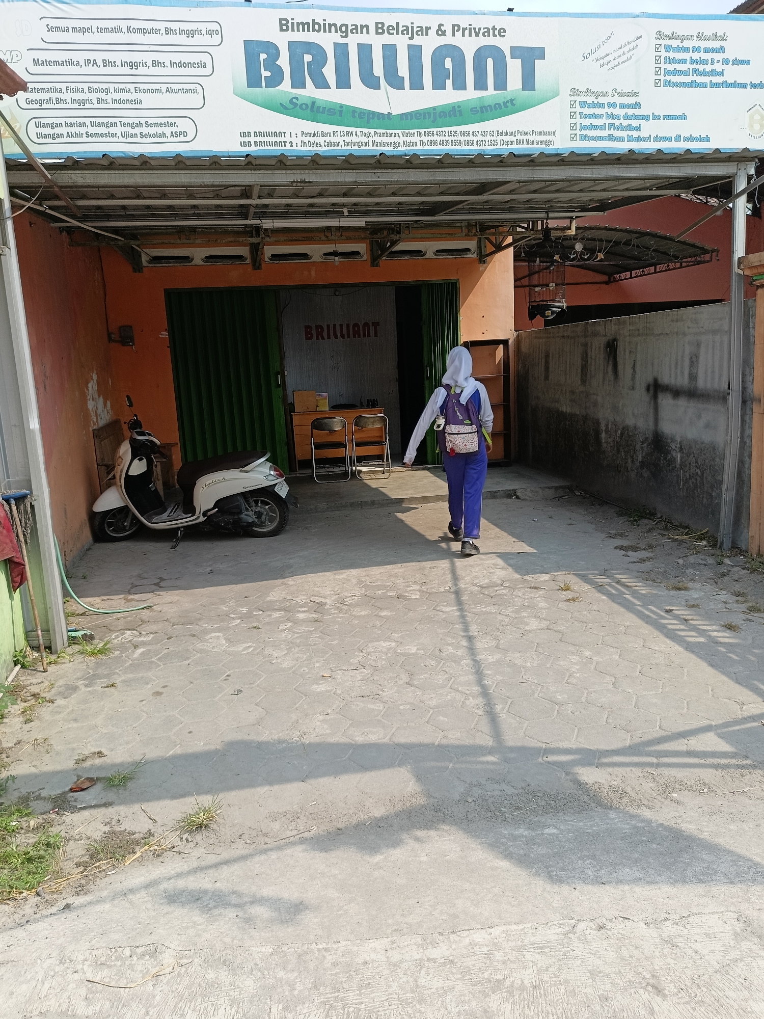 A young person wearing a white hoodie and blue pants, with a backpack, walks away from the camera down a paved path in front of a commercial building. The building has an orange facade with a corrugated metal roof and a green roll-up door. A white scooter is parked to the left of the building entrance. A sign above the entrance reads "Bimbingan Belajar & Private BRILLIANT Solusi tepat menjadi smart" and lists phone numbers and addresses in Manisrenggo, Klaten, Indonesia. The ground is paved with hexagonal concrete tiles and there is some vegetation and debris present. The scene is lit by sunlight, casting shadows on the ground.