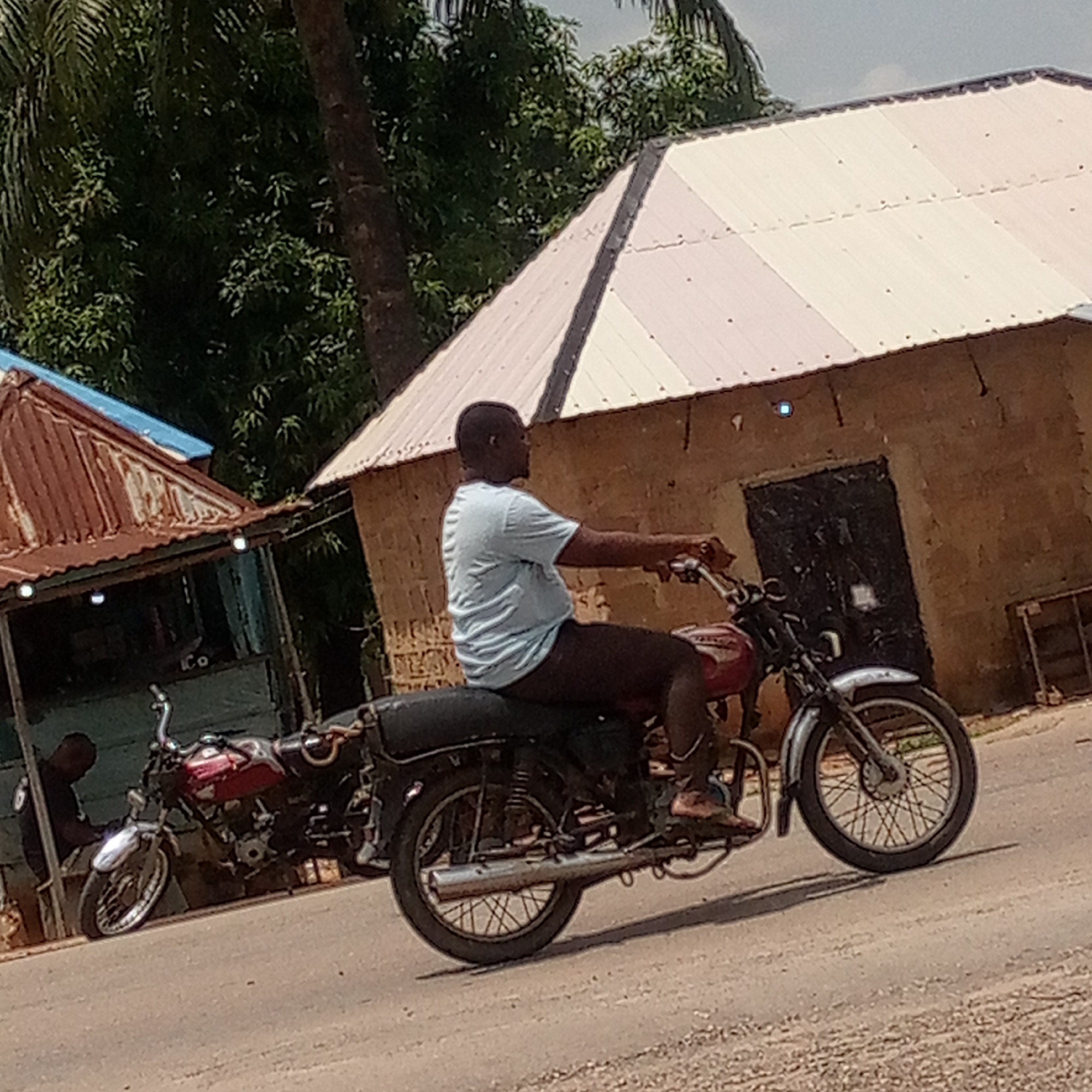 The image captures a daytime scene in what appears to be a semi-urban or rural setting in Mararaba, Nigeria. The central subject is a man riding a red and black motorcycle on a light-colored, possibly paved and somewhat dusty, road. He is wearing a light blue t-shirt and dark trousers, with his bare feet visible, one resting on the motorcycle's footrest. He is viewed from the side, looking towards the left of the frame.
In the background, two simple buildings are visible. To the left, a structure with a rusty, corrugated iron roof and a blue section above it stands next to another parked motorcycle. A person, largely obscured, is visible near this parked motorcycle. To the right, a larger building features light-colored, possibly white or light grey, corrugated iron roofing and mud-colored walls, with a dark door. Lush green foliage and tree trunks are prominent in the background, suggesting a verdant environment. The sky is bright, indicating clear weather. No specific text is clearly visible in the image. The overall impression is a snapshot of everyday life and transport in the described location.