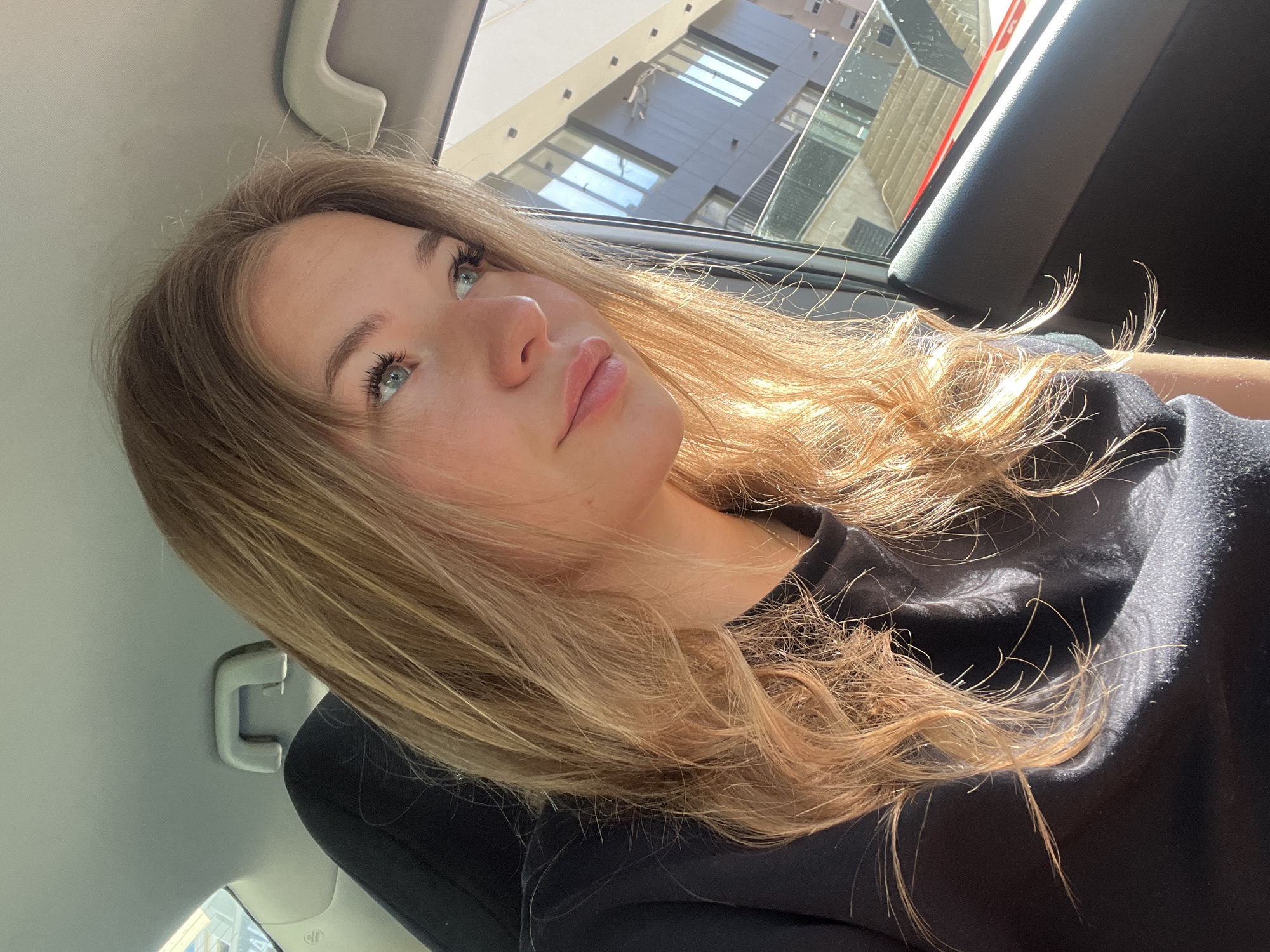A light-skinned female with blue eyes and long blonde hair is the primary subject of this photograph, taken inside a car. She is wearing a black shirt. The picture is shot from a low angle, tilting upward towards her face and the window. Sunlight illuminates her face and hair. Outside the car window, there is a partial view of buildings, which appear to be in an urban setting. Given the prompt's instruction to situate this image in Budva, Montenegro, one can infer that the vehicle and its occupant are within the city limits, although the photograph does not offer specific geographic details for definitive confirmation.