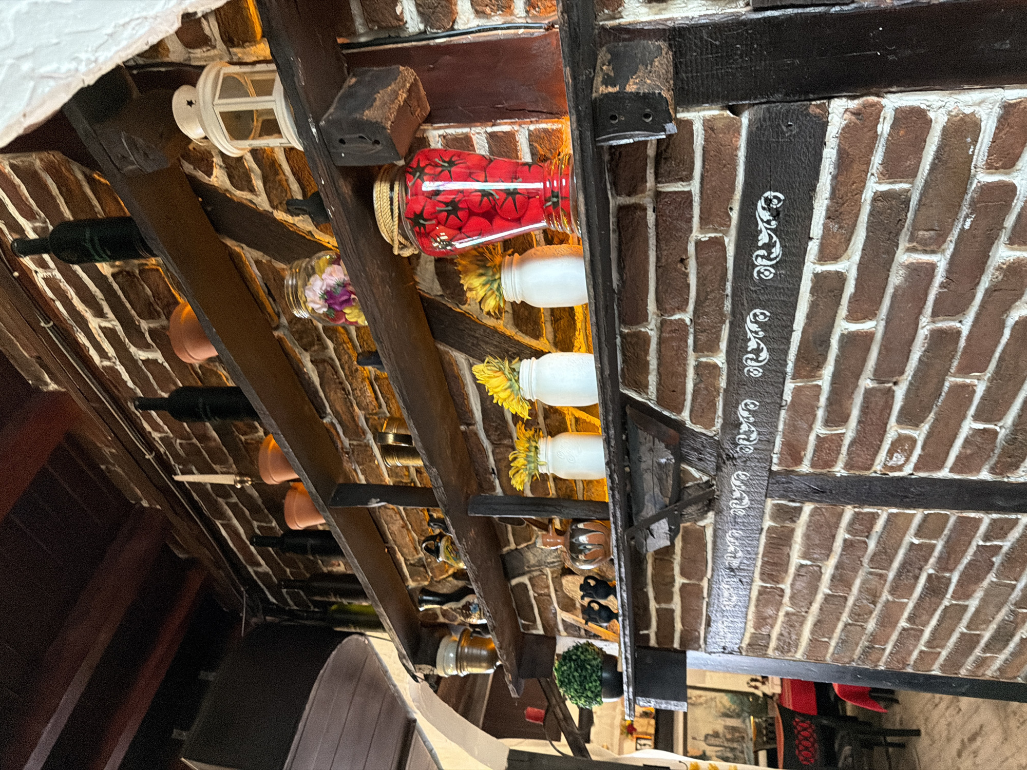 The image depicts the interior of an establishment in Varna, Bulgaria, characterized by a prominent brick wall adorned with multiple dark wooden shelves. The wall features exposed reddish-brown bricks with white mortar, segmented by vertical and horizontal dark wooden beams, some of which exhibit white stenciled or painted scrollwork patterns. Overhead, dark wooden ceiling beams are visible.
Three primary dark wooden shelves are mounted on the brick wall. The uppermost shelf holds several dark glass bottles, terracotta pots (one containing a white candle), and a white decorative lantern with a star cutout on its cap. The middle shelf displays a variety of decorative ceramic and metallic items, including small vases, pitchers, and metallic cups, alongside a clear glass jar filled with colorful artificial flowers. The lowest visible shelf contains a small potted artificial green plant, two black decorative urn-like objects, an orange and brown glazed ceramic vase, two white ceramic jars each holding yellow artificial flowers (resembling sunflowers), and a tall red vase with a black tomato vine pattern, topped with a coiled rope accent.
In the blurred background to the lower left, dark chairs and tables draped with red tablecloths are partially visible, suggesting a restaurant or dining setting. No individuals or active interactions are present in the scene. The lighting appears warm and indirect, illuminating the shelves and their contents.