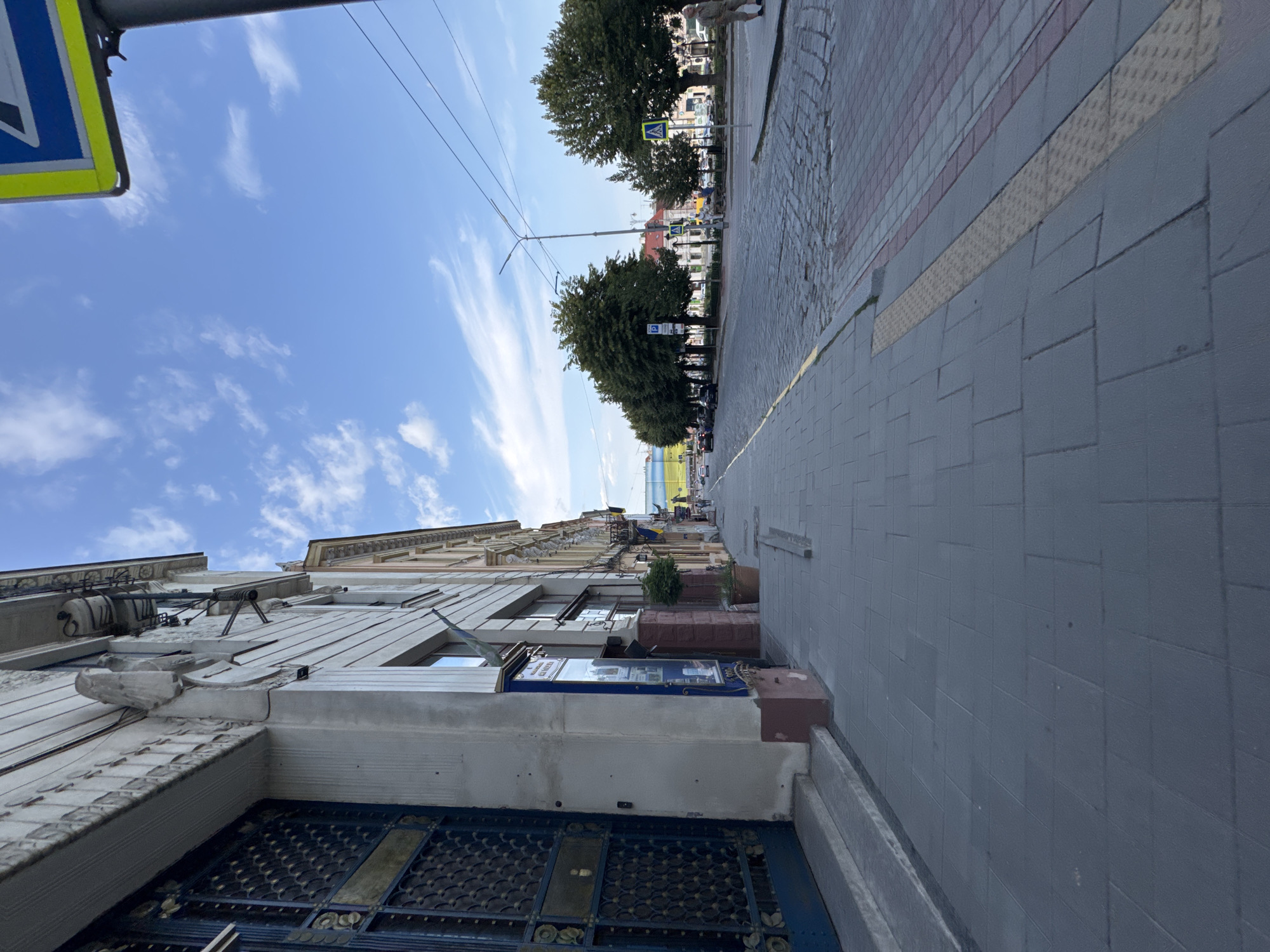 The image captures a street scene in Chernivtsi, Ukraine, on a cloudy day. The perspective is from a low angle, looking down a paved sidewalk lined with buildings on the left and a cobblestone street on the right. The architecture of the buildings appears to be old and ornate, with decorative facades and large, dark-colored metal-framed doors or windows. Trees line the opposite side of the street, with a few cars and pedestrians visible in the distance. A crosswalk sign is prominent in the upper right corner of the image. The overall atmosphere is that of a typical European city street.