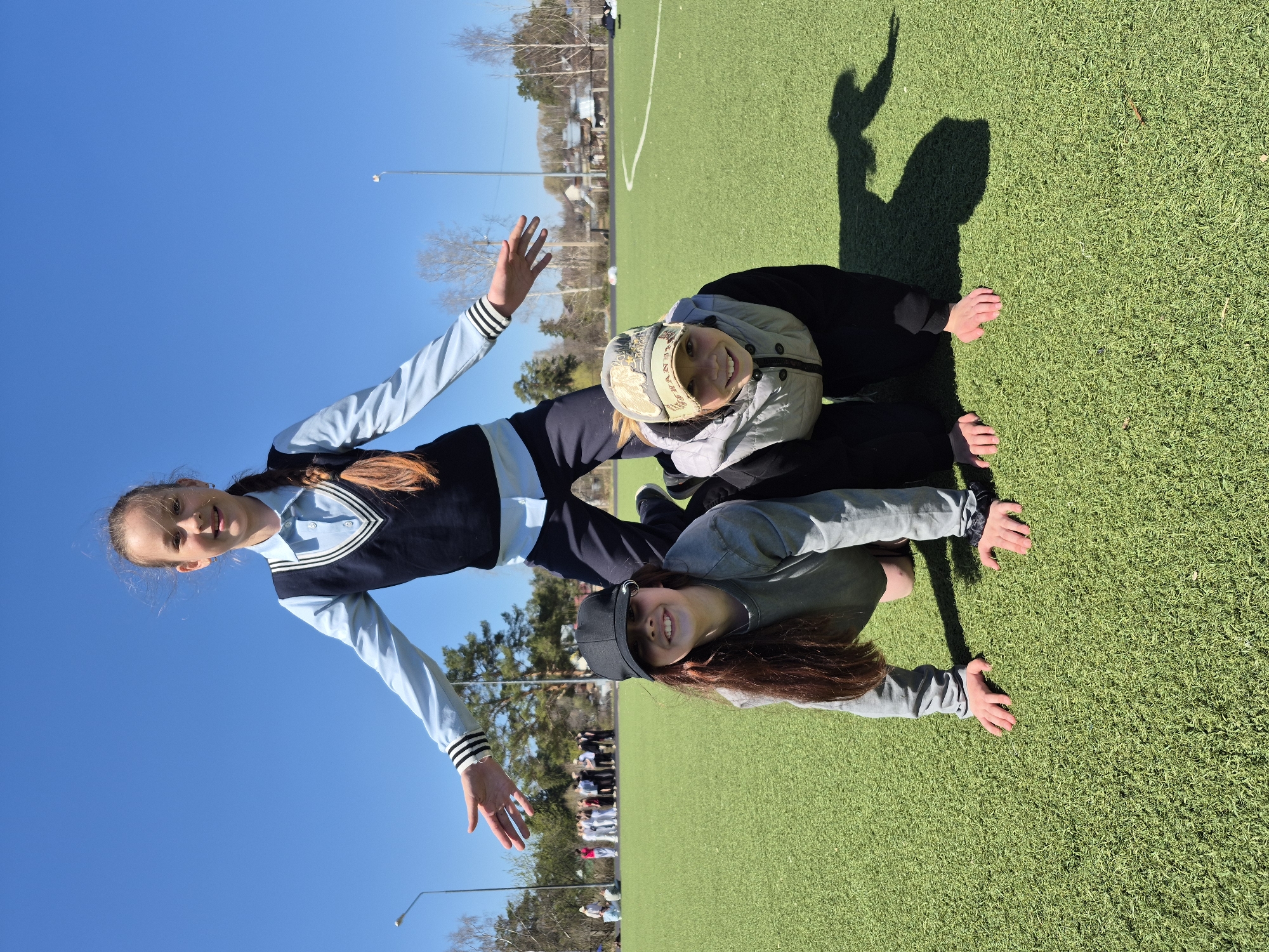 Three girls are arranged in a stacked position on a green artificial turf field, likely a sports field or playground. The top girl is wearing a light blue school-style top with a dark blue vest and has her arms outstretched towards the sky. The middle girl is wearing a khaki hat and grey jacket. The bottom girl is wearing a grey jacket and black cap. They are situated in front of a clear blue sky. In the background, there's a row of people near a line of trees and buildings. Considering the specification of Chaikovsky, Russia, the location might be a public park or school grounds featuring a sports field in that city.