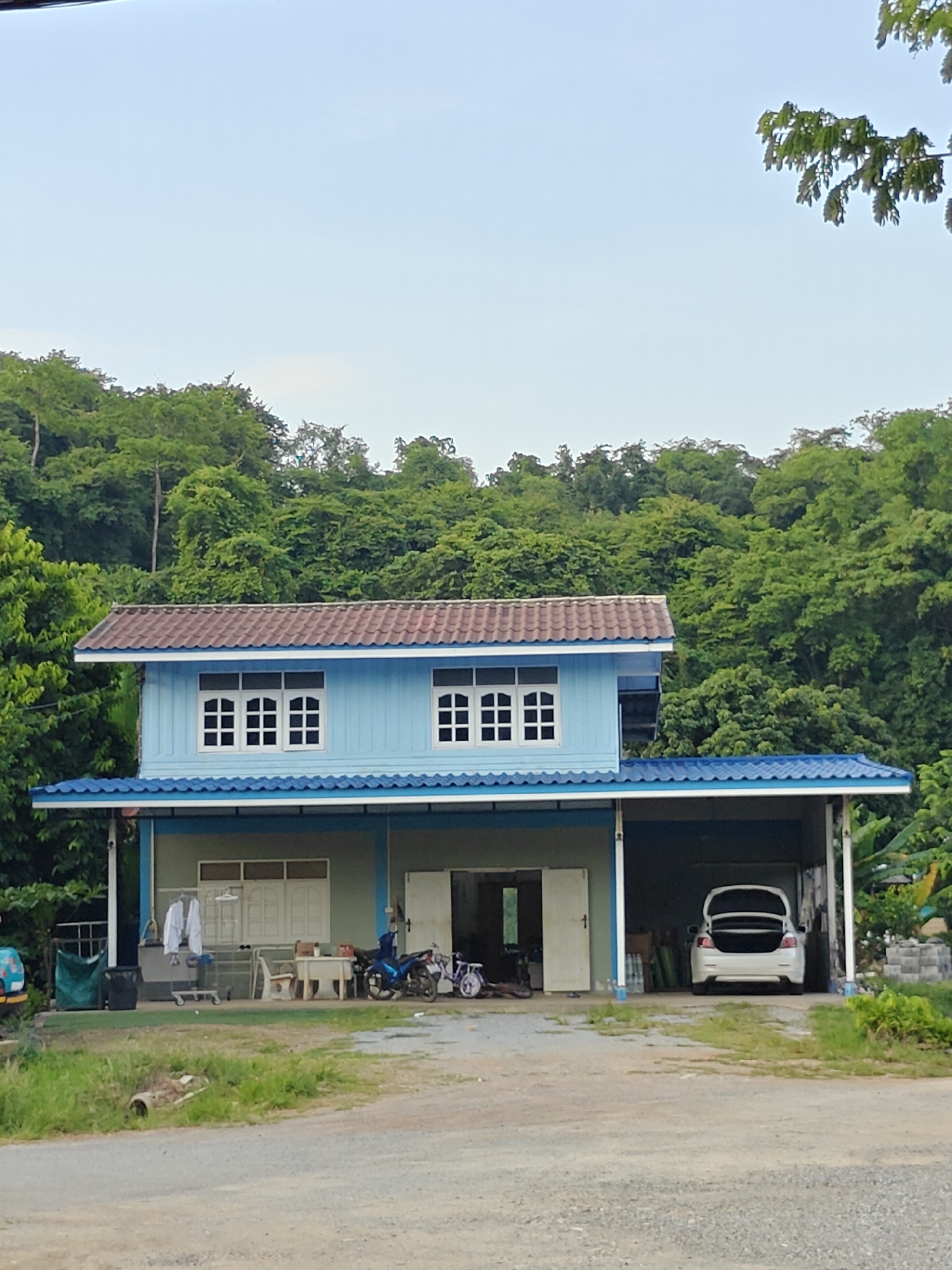 The image shows a two-story light blue house with a brown tiled roof, located in Mu Si, Thailand. The house has white-framed windows on the second story and a blue-tiled overhang on the first story. The first floor features an open carport with a white car parked inside, its trunk open. To the left of the carport, there are doors and windows, with laundry hanging on a rack outside. A motorcycle is parked in front of the house. The house is situated in a natural environment with lush green trees and foliage in the background, and a dirt and gravel road in the foreground. The sky is clear and light blue.