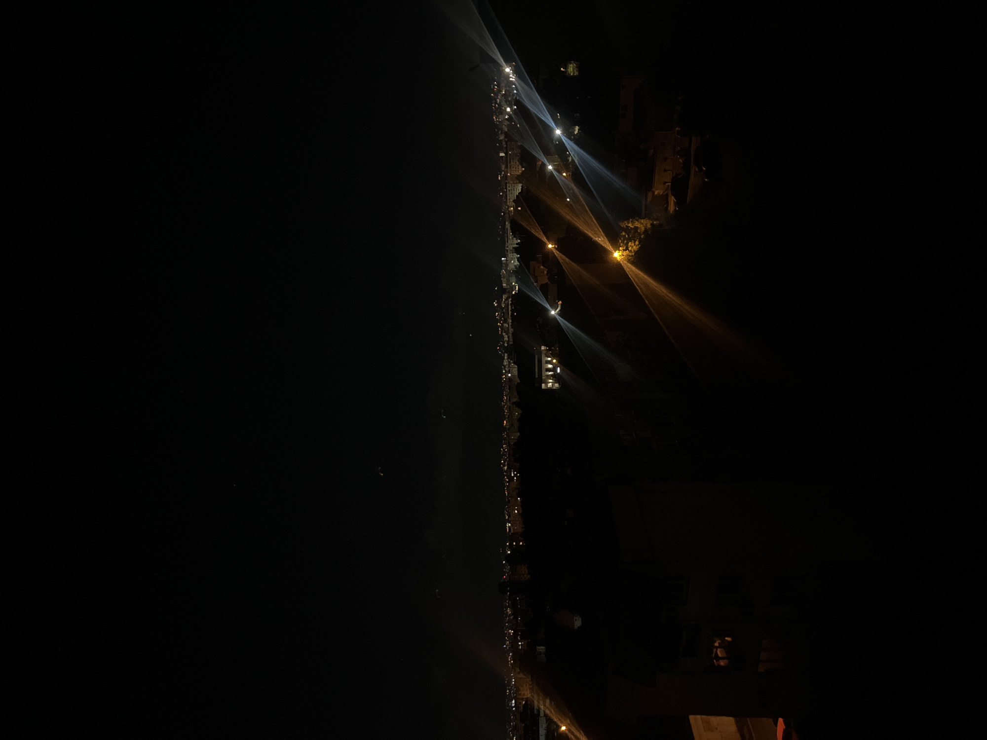 An aerial, dark view of a street illuminated by streetlights, potentially along the coastline of Sofia, Bulgaria. The street lights cast long beams of light onto the dark surface to the left. Buildings of varying heights and orientations are visible on the right. The sky is dark.