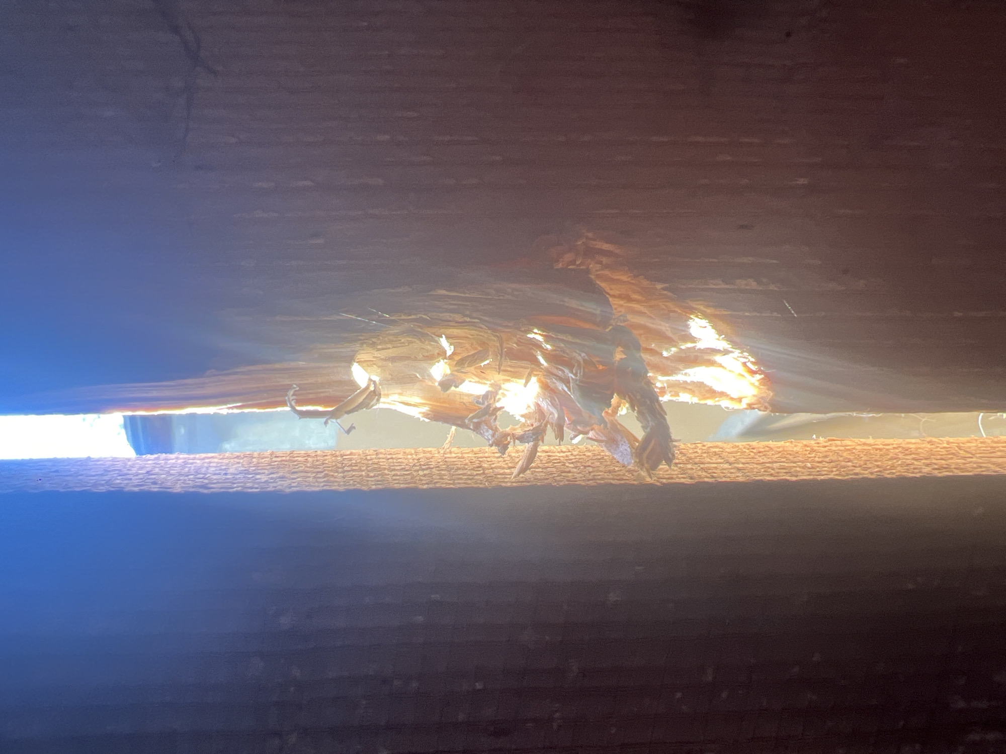 The image displays a close-up view of damaged wood, likely from a structural element like a ceiling or wall. The wood is splintered and broken in the center, revealing a gap through which light is streaming, creating a dramatic effect with sunbeams. The texture of the damaged wood appears rough and fibrous.
Below the damaged section, there is a horizontal surface with a textured pattern, possibly another wooden plank or beam. This surface is illuminated by the light filtering through the damage above, creating subtle shadows and highlights. The area below this textured surface is darker, with horizontal lines suggesting the grain or construction of another wooden component.
The lighting suggests it is daytime, with sunlight actively penetrating the opening. The scene is focused on the structural damage and the interplay of light and shadow within this confined space. No people, animals, or specific objects are clearly identifiable beyond the wooden elements. The overall impression is one of decay or damage to a wooden structure, with the light emphasizing the breakdown. The location is not identifiable from the visual information provided. There is no visible text in the image.